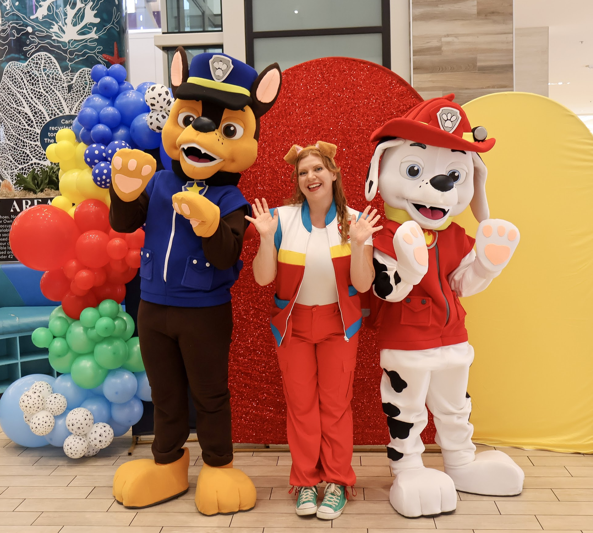 Two costumed dog mascots and a host at a colorful themed mall photo area