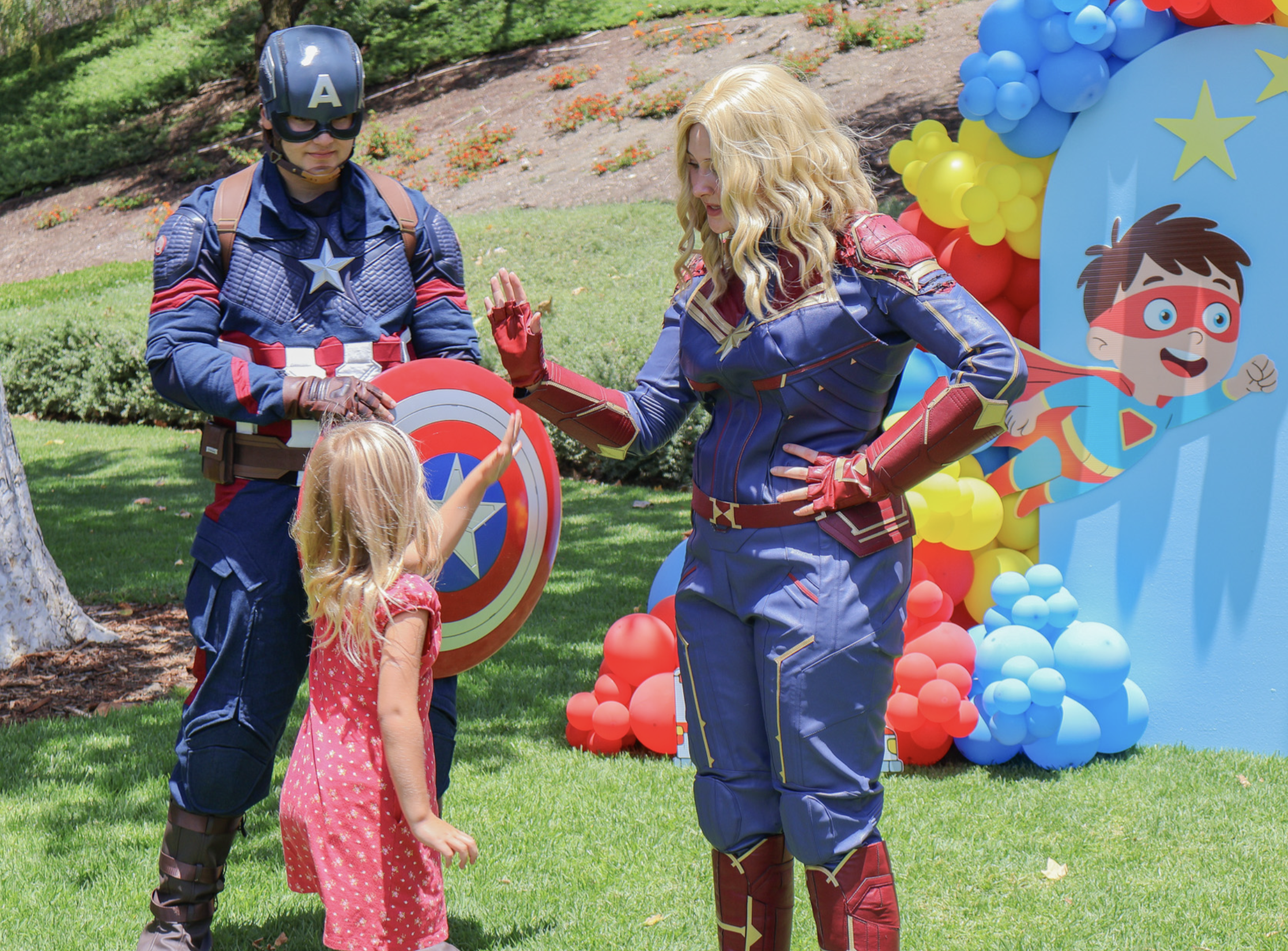 Captain America and Captain Marvel interacting with a young guest beside a superhero photo backdrop