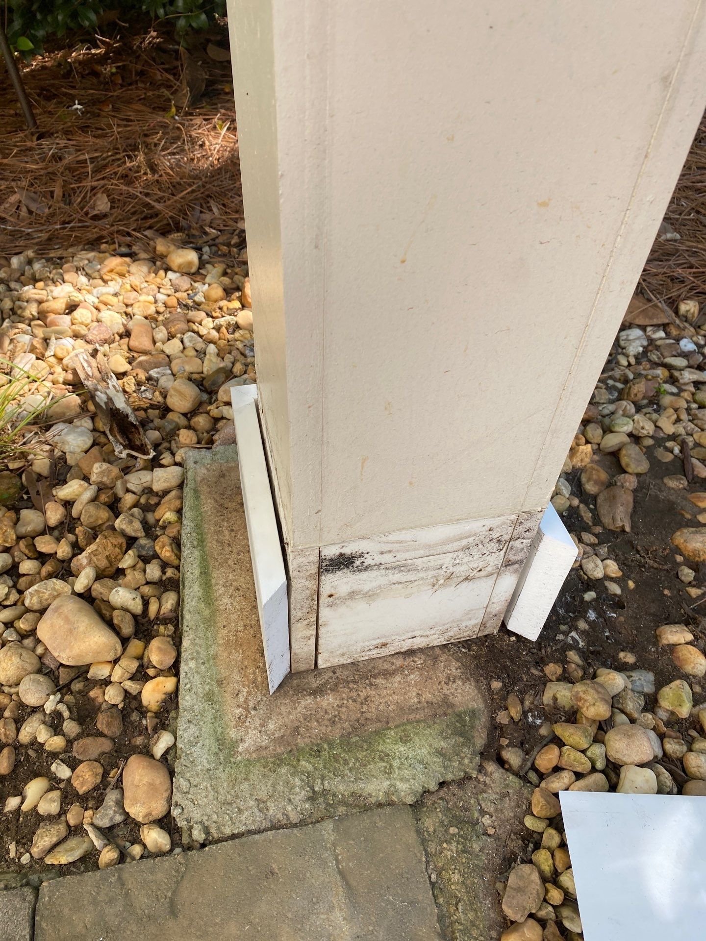 Trim board at the base of a column with a dark rotted section against landscape rock