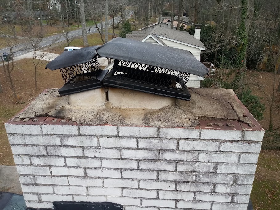 Black chimney cap with mesh sides seated on a white brick chimney crown, houses and trees behind
