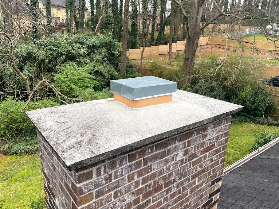 Brick chimney with a concrete mortar crown and a metal chase cap, seen from the roof