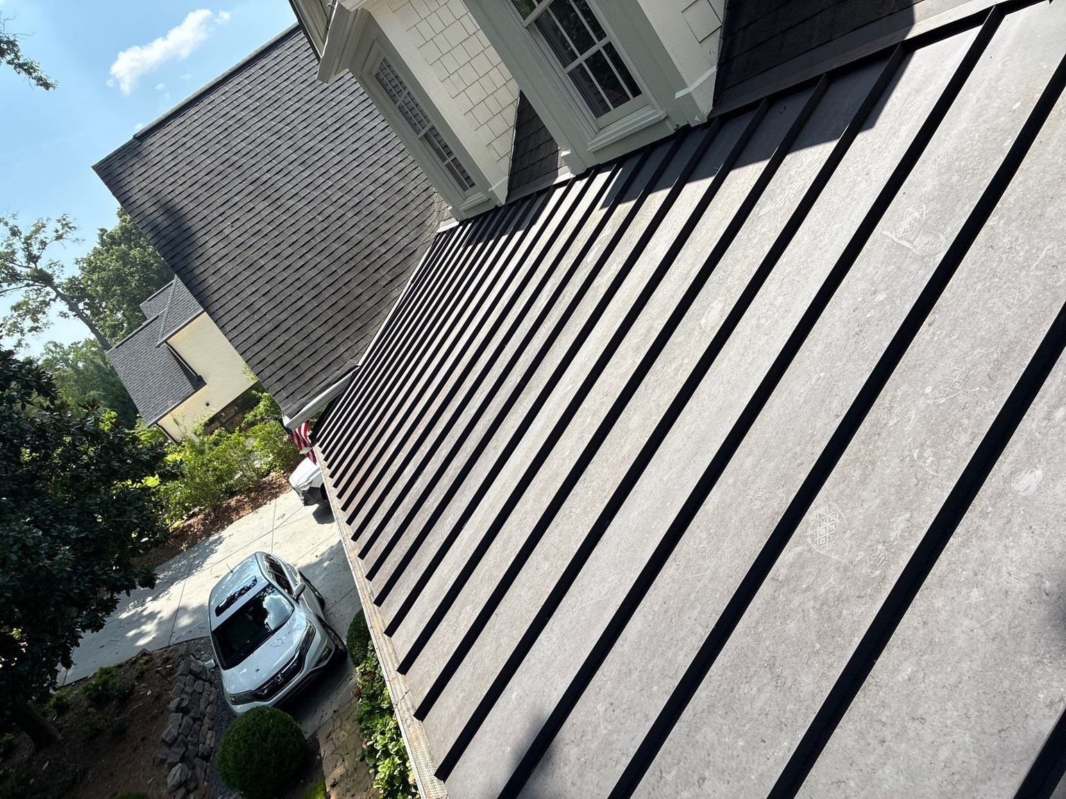 Dark standing seam metal roof panels running down toward a gutter line above a brick wall and driveway