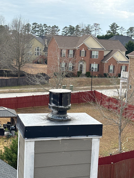 Black vent cap seated on a chimney chase above vinyl siding, neighborhood in the background