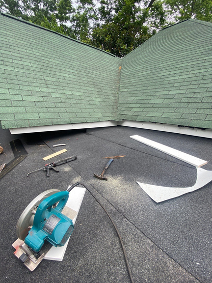 Flat roof work in progress with a circular saw, new membrane, and flashing pieces next to a green shingle wall