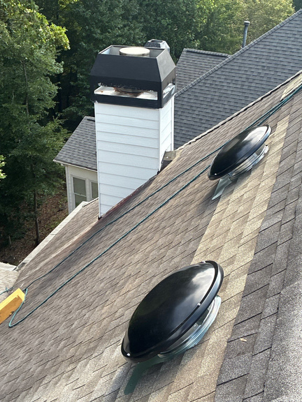 Two dome style powered attic fans on a brown shingle roof beside a white sided chimney chase