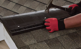 Worker in a red glove fastening ridge vent material along a shingle roof peak