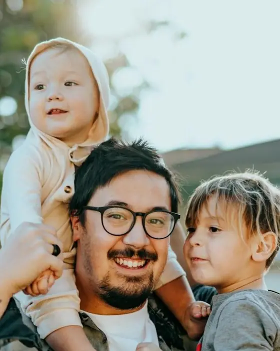 A smiling father carries his baby on his shoulders next to his son
