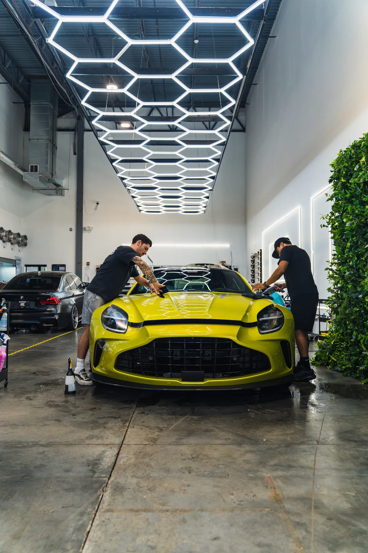 Two technicians finishing a yellow Aston Martin Vantage under the hexagonal light ceiling