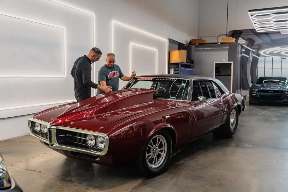 A client and a technician talking over a red Pontiac Firebird