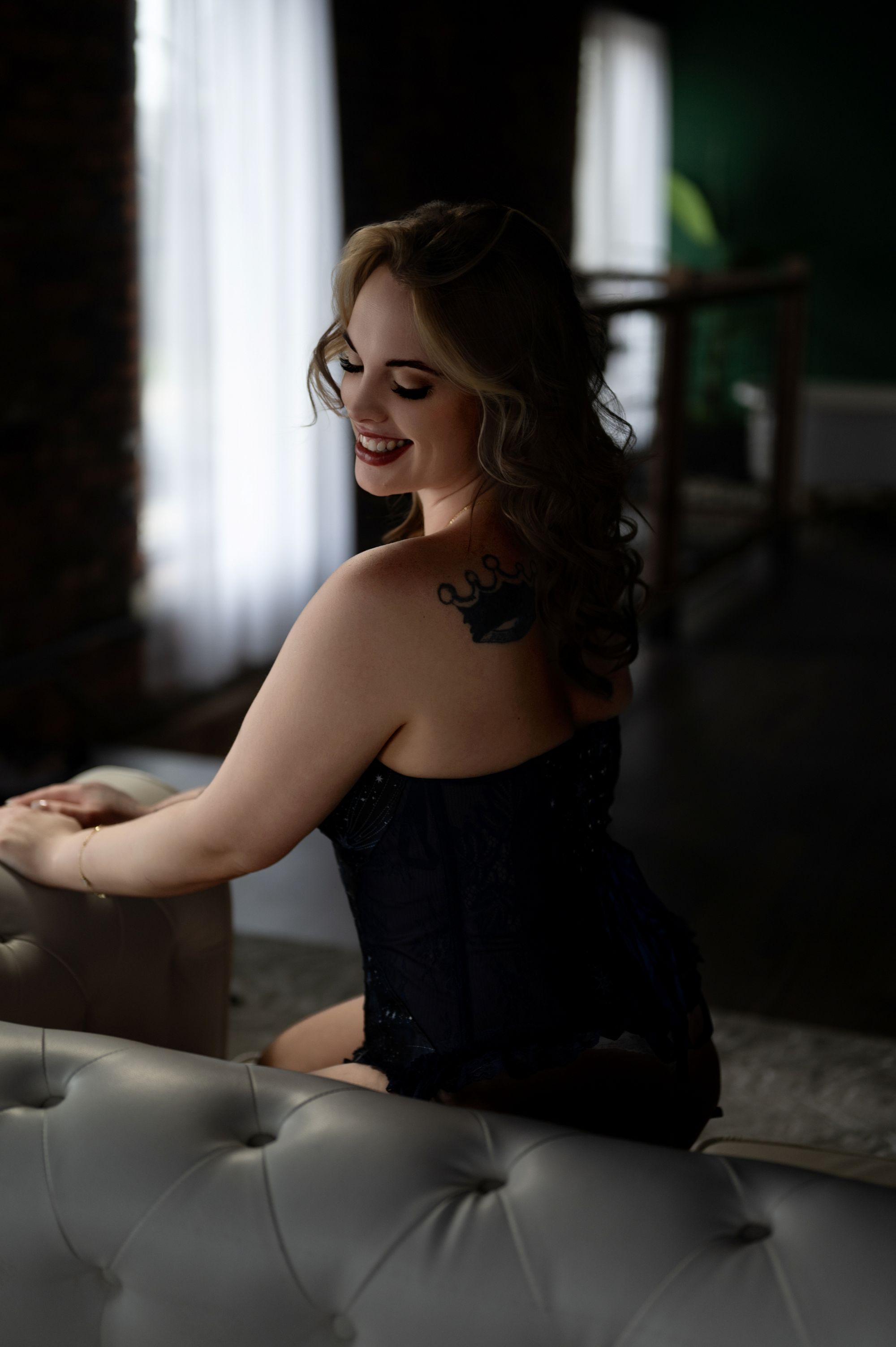 Smiling woman in a black corset sitting on a tufted sofa with natural light and exposed brick backdrop – elegant boudoir session.