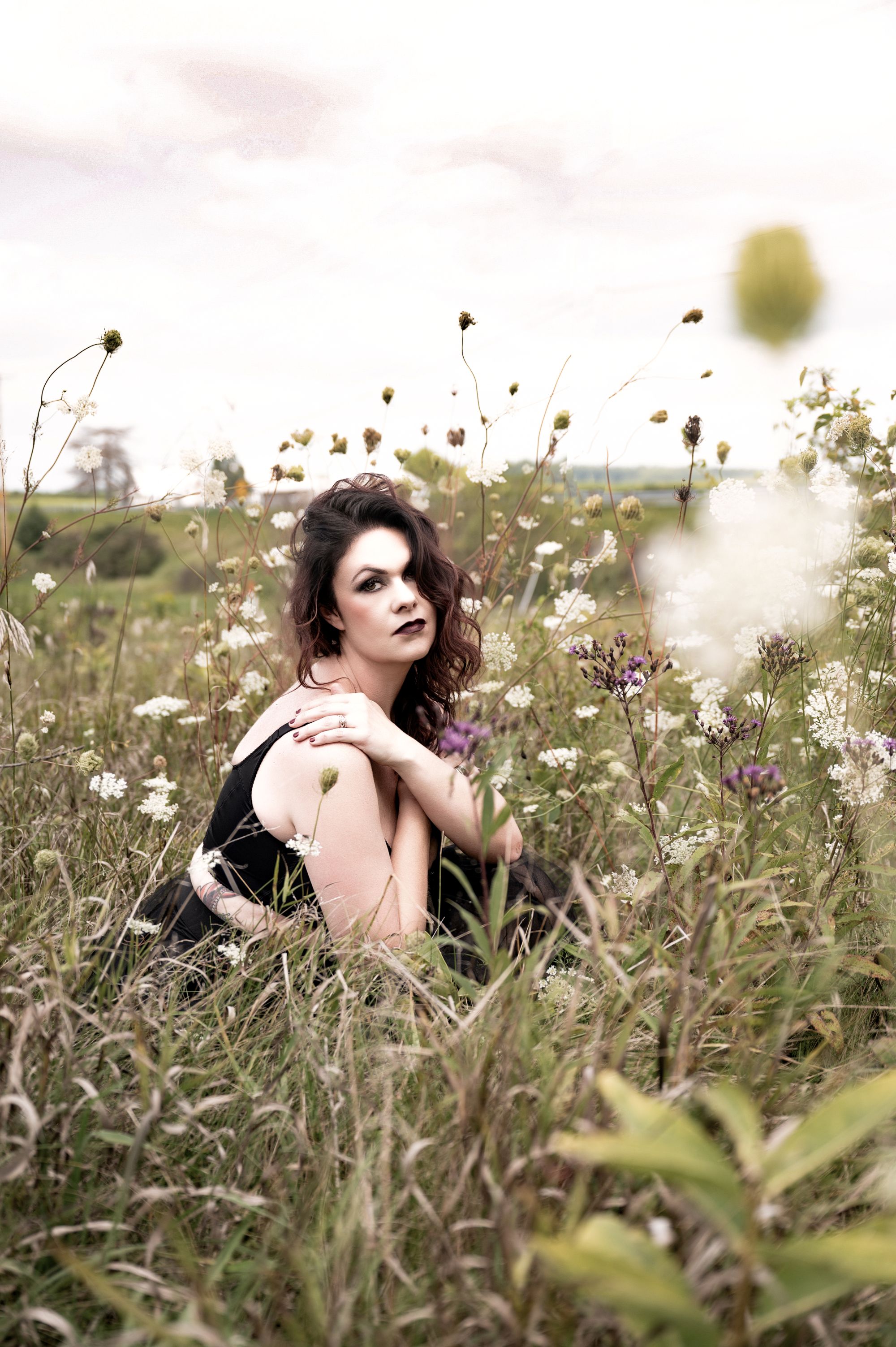 Moody outdoor portrait of a woman in a black dress sitting among tall grass and wildflowers with a dramatic sky overhead.