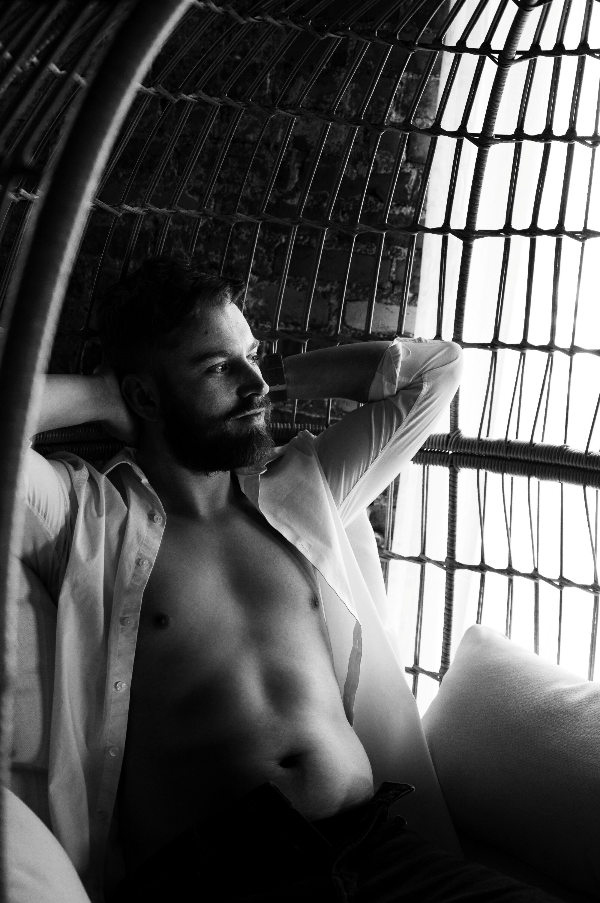 Black and white male boudoir portrait of a man reclining shirtless in a wicker chair, highlighting masculine confidence and sensuality.