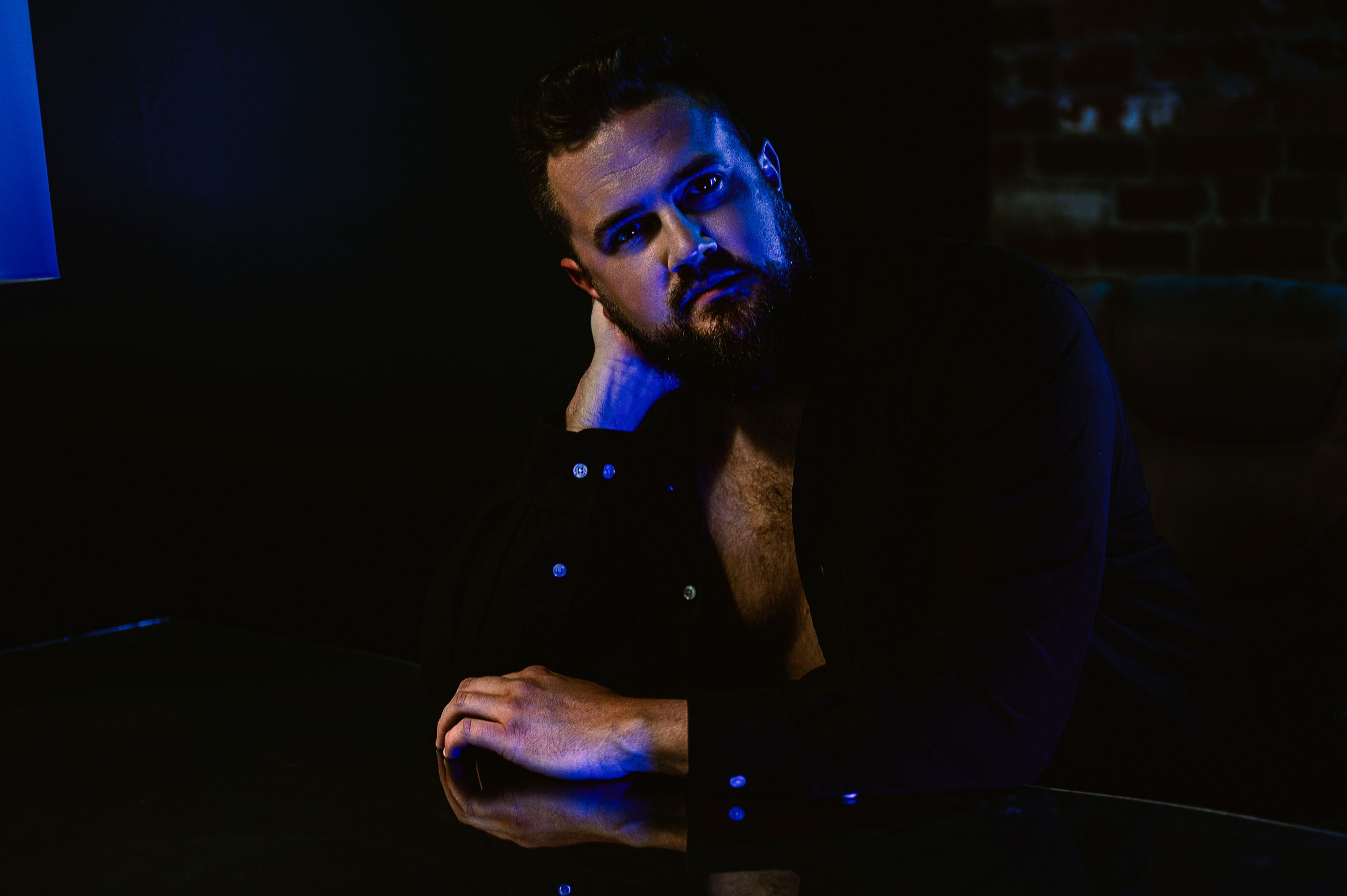 Moody male boudoir portrait of a man in an open black shirt with dramatic blue lighting, capturing masculine strength and allure.