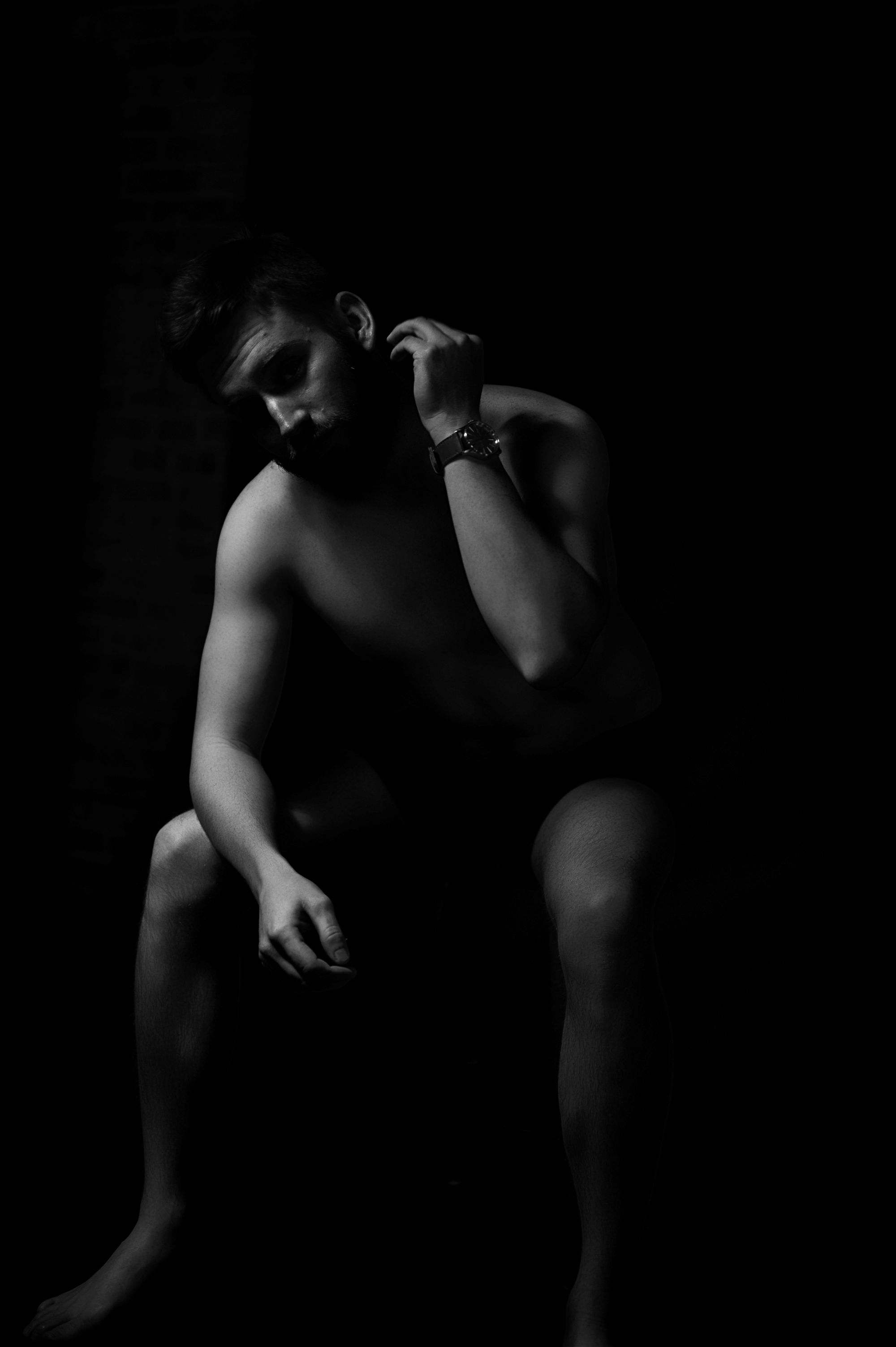 Dramatic black and white boudoir portrait of a man sitting shirtless in shadows, emphasizing strong masculine form and moody elegance.