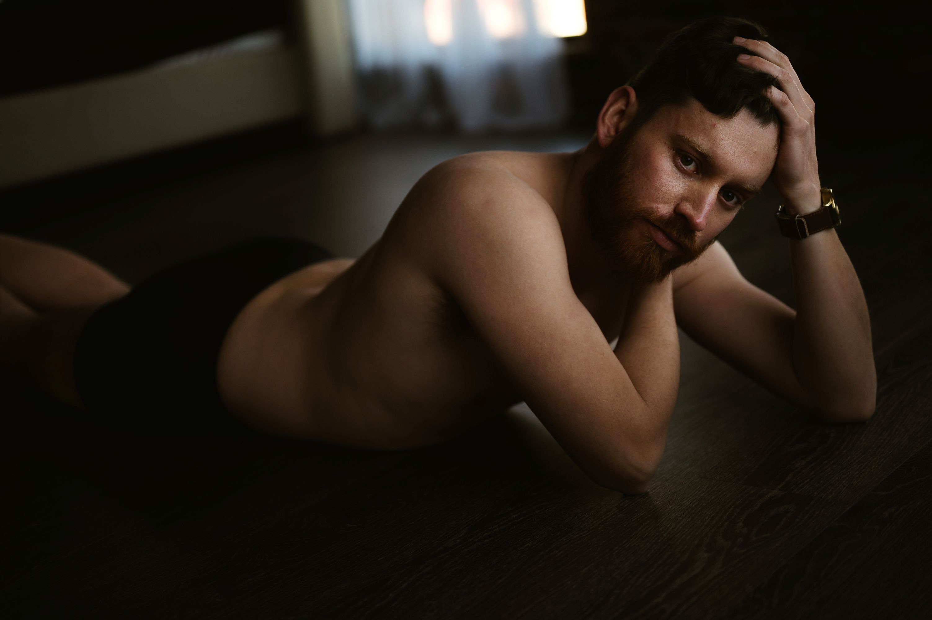 Artistic male boudoir portrait of a shirtless man lying on the floor, posing with a pensive gaze and confident expression.