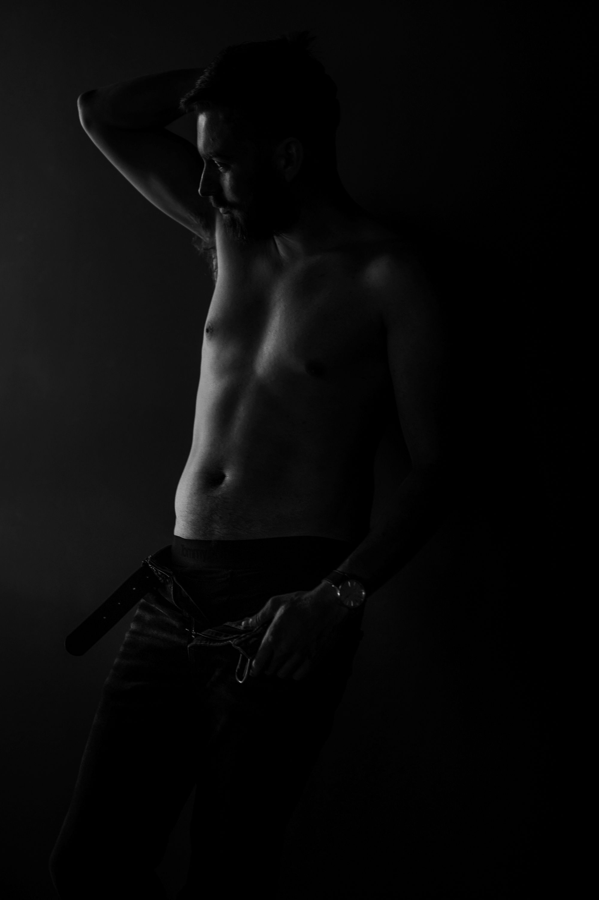 Dramatic male boudoir photo of a shirtless man with undone belt, captured in moody lighting to emphasize masculine sensuality.