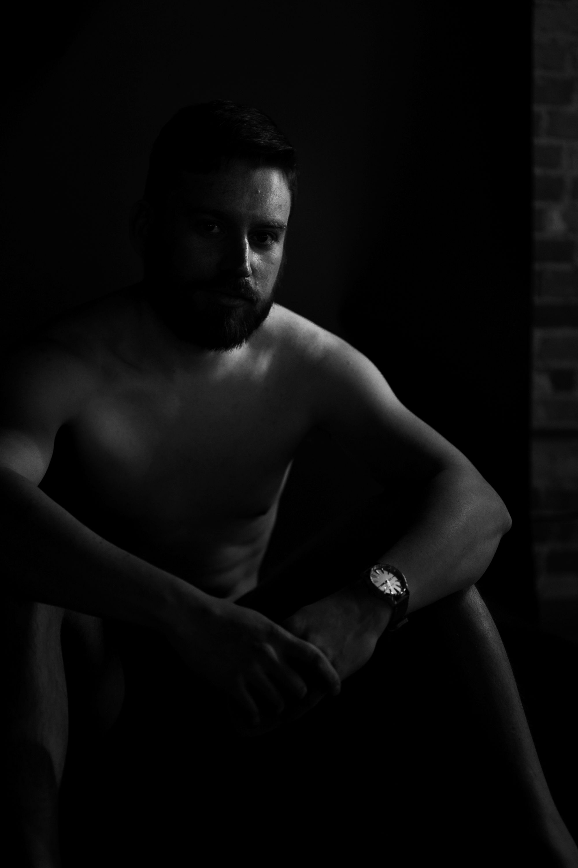 Black and white male boudoir portrait of a shirtless man sitting in shadows, highlighting confidence and strong masculine features.