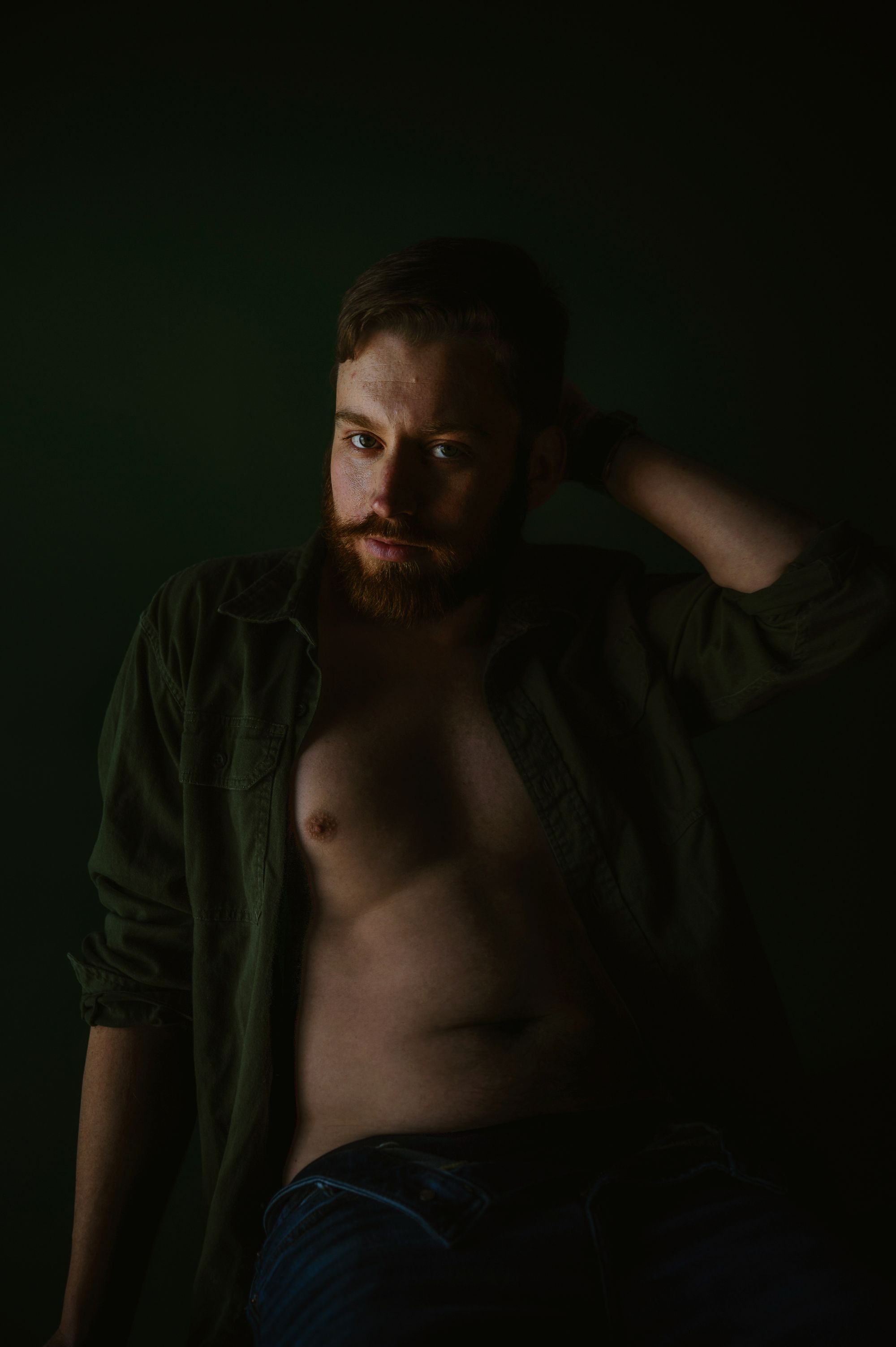 Male boudoir photo of a man with beard and open shirt against dark green backdrop, embracing raw masculine confidence.