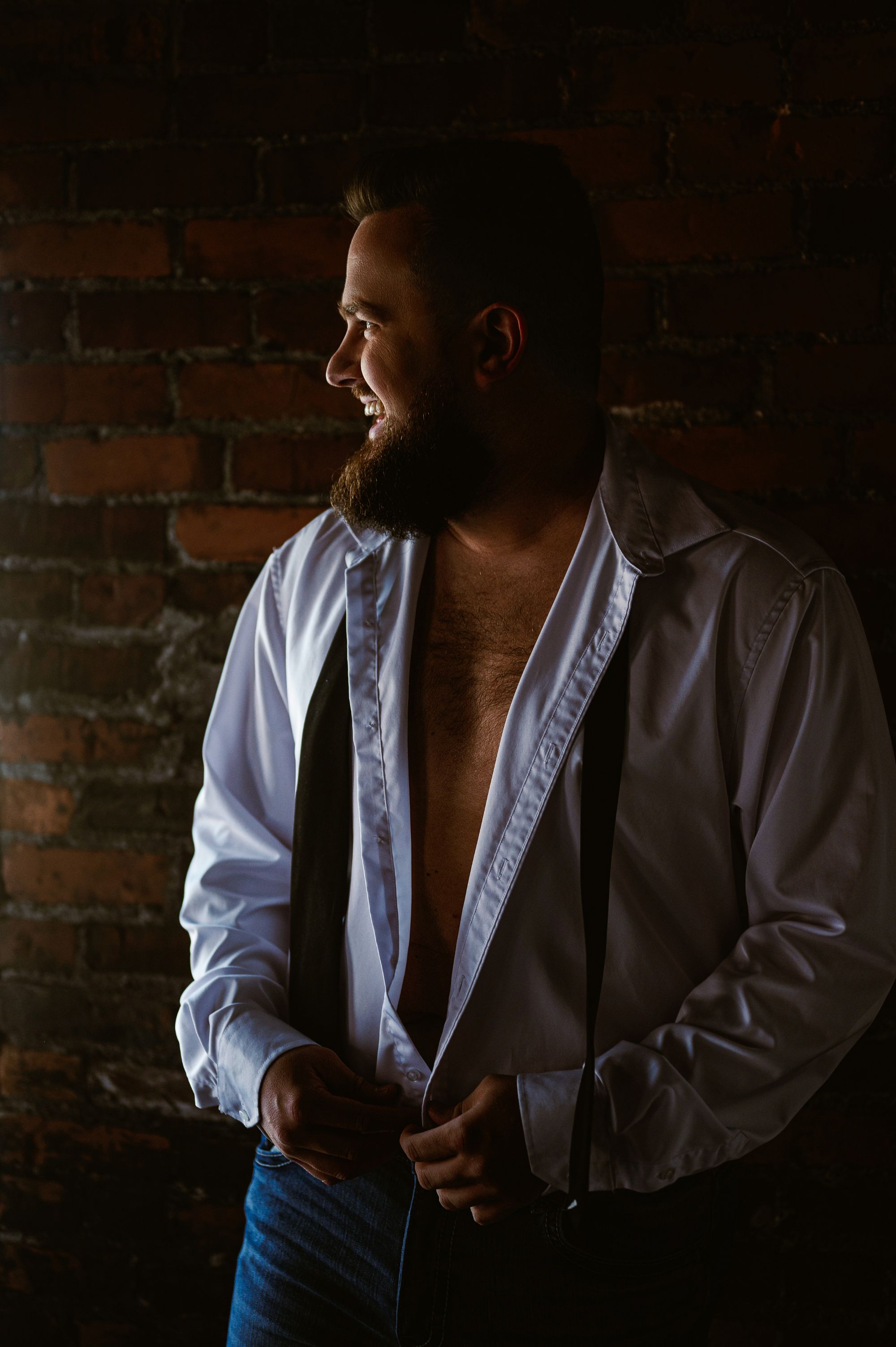 Playful male boudoir portrait of a man smiling in an open white shirt with suspenders, captured against a rustic brick wall.