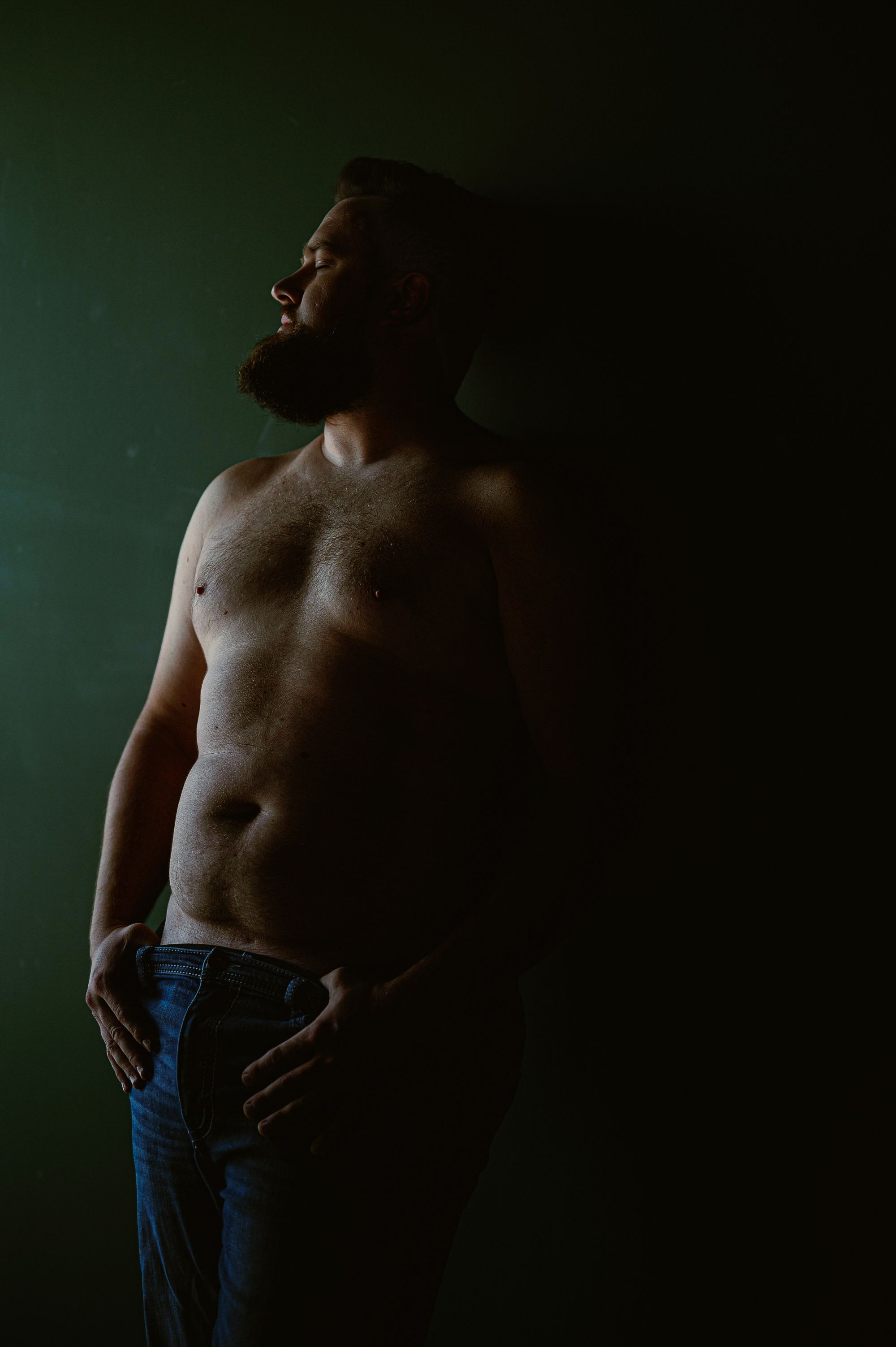 Intimate male boudoir photography of a shirtless man in jeans leaning against a wall with eyes closed, exuding strength and vulnerability.