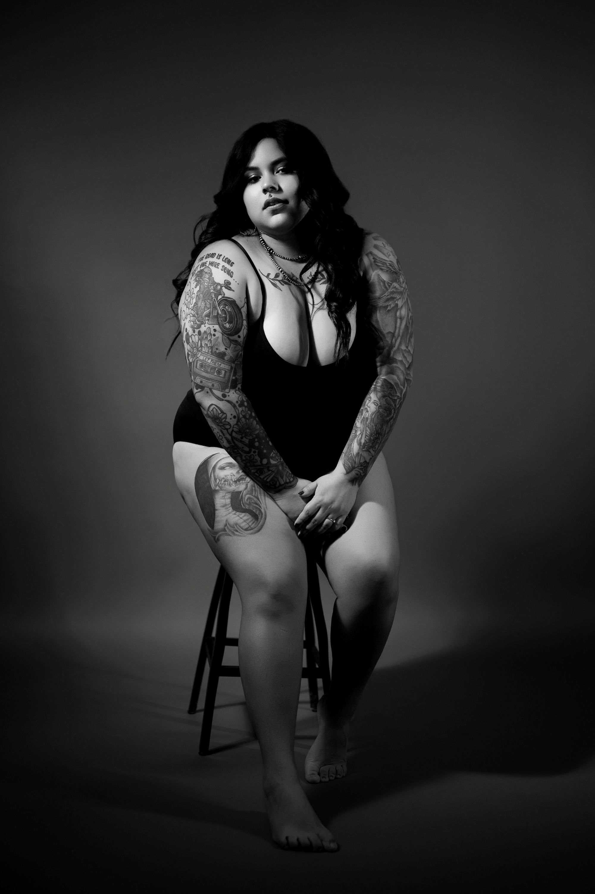 Bold boudoir portrait of a tattooed woman in a black bodysuit sitting on a stool, embracing confidence in studio lighting.