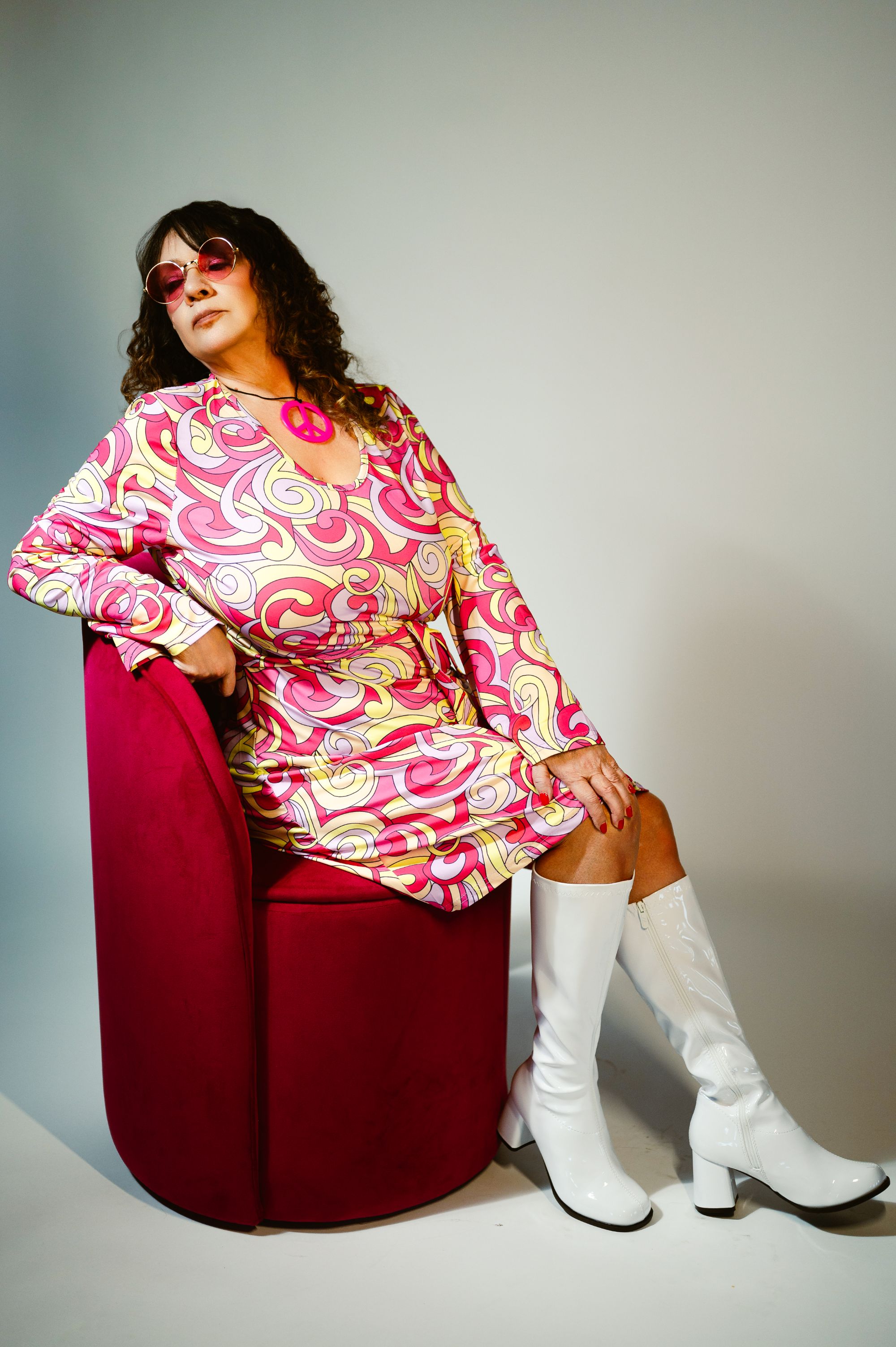 Retro fashion portrait of a woman in a colorful 70s dress with white go-go boots and a peace sign necklace.