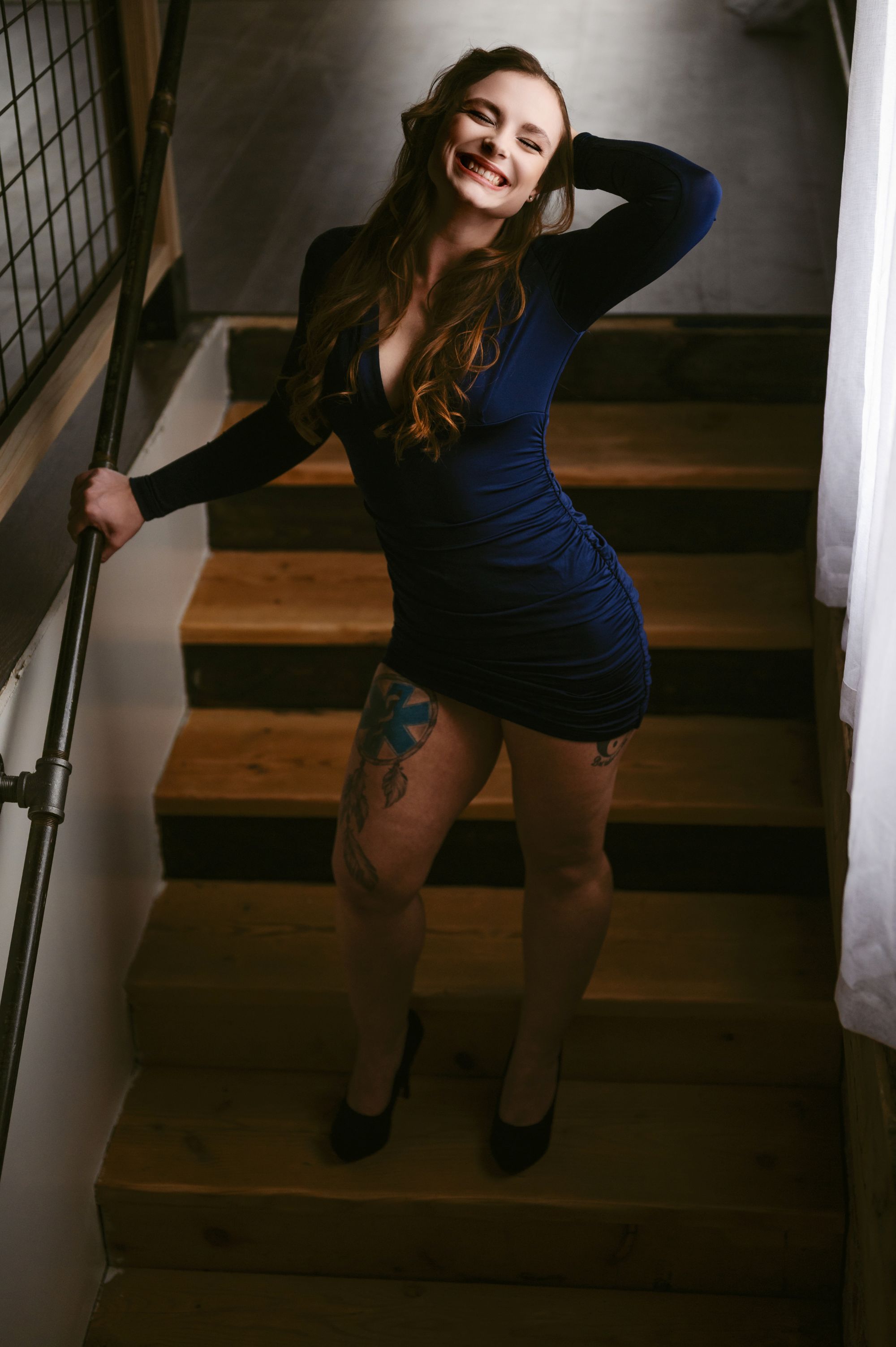 Happy woman in a fitted navy blue dress posing on a staircase with a bright, joyful smile.