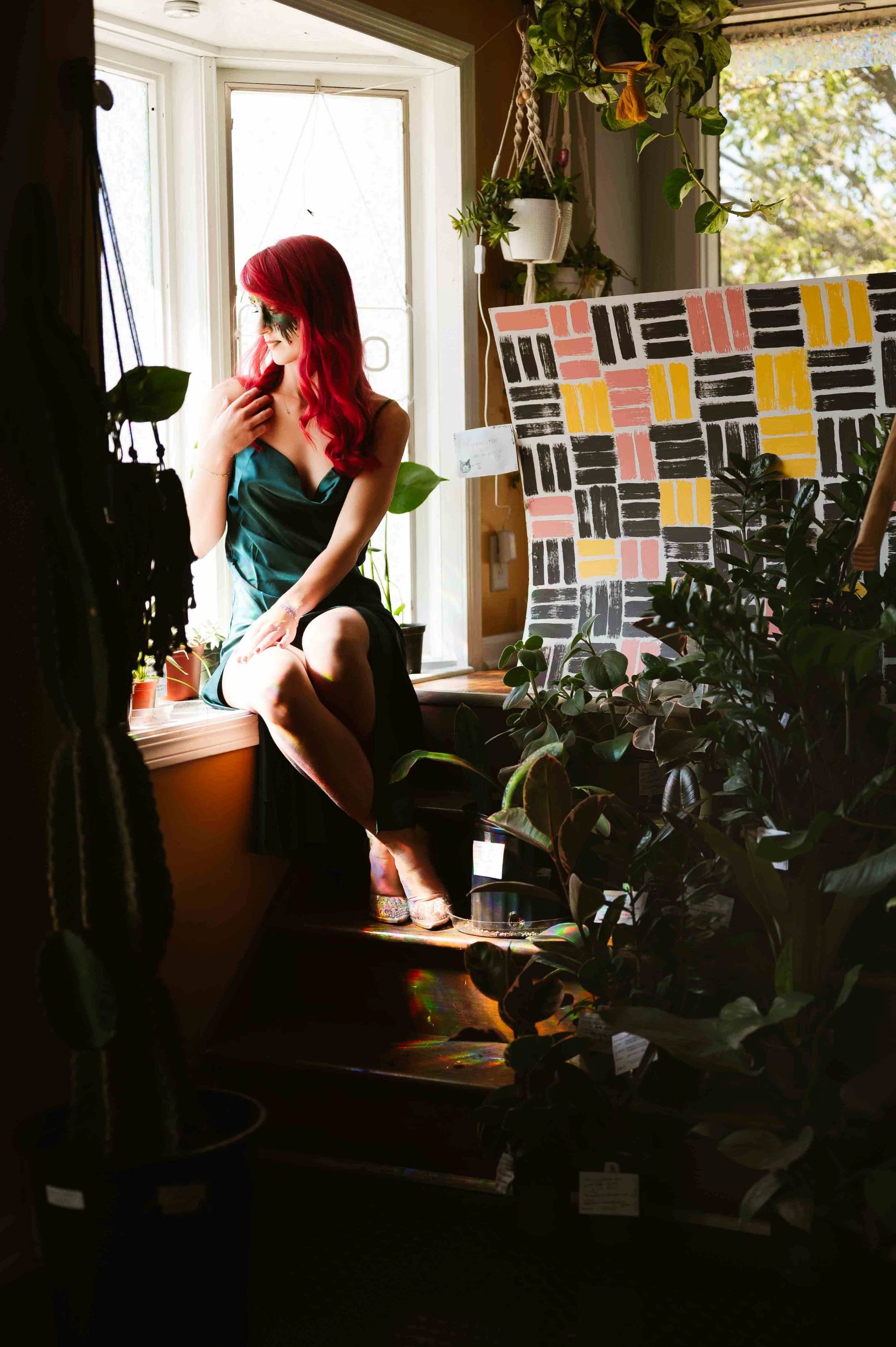 Portrait of a red-haired woman in a green dress sitting by a window surrounded by lush indoor plants