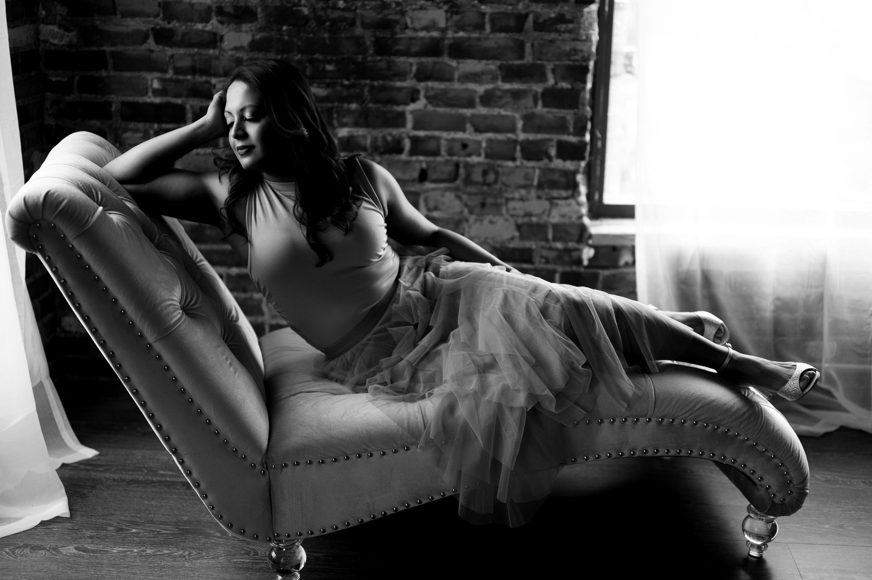 Black and white portrait of a woman reclining gracefully on a tufted chaise lounge in front of a brick wall and window light.