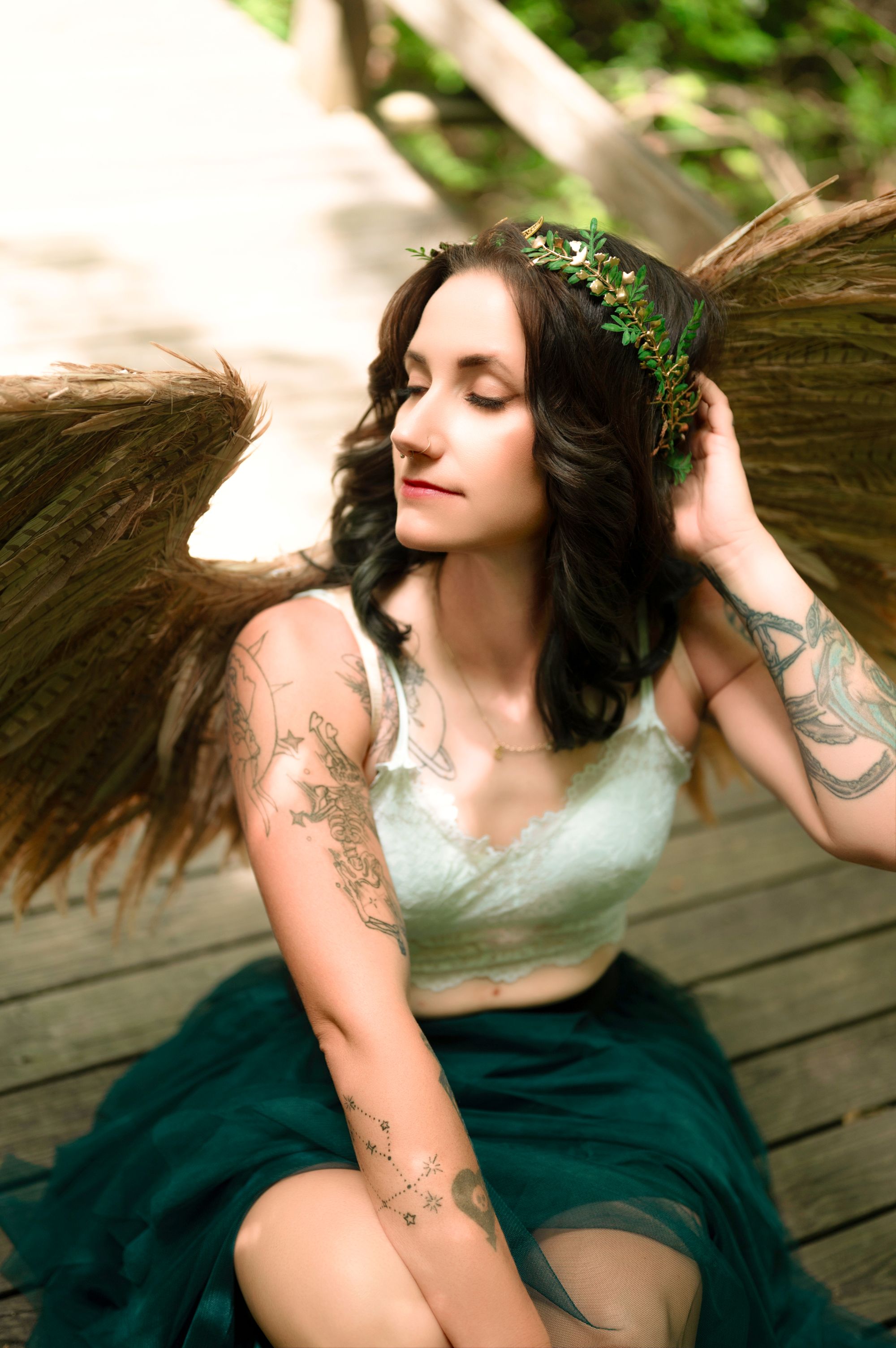Fantasy-inspired portrait of a tattooed woman wearing feathered angel wings, a green skirt, and a crown-like headpiece while sitting on a wooden bridge.