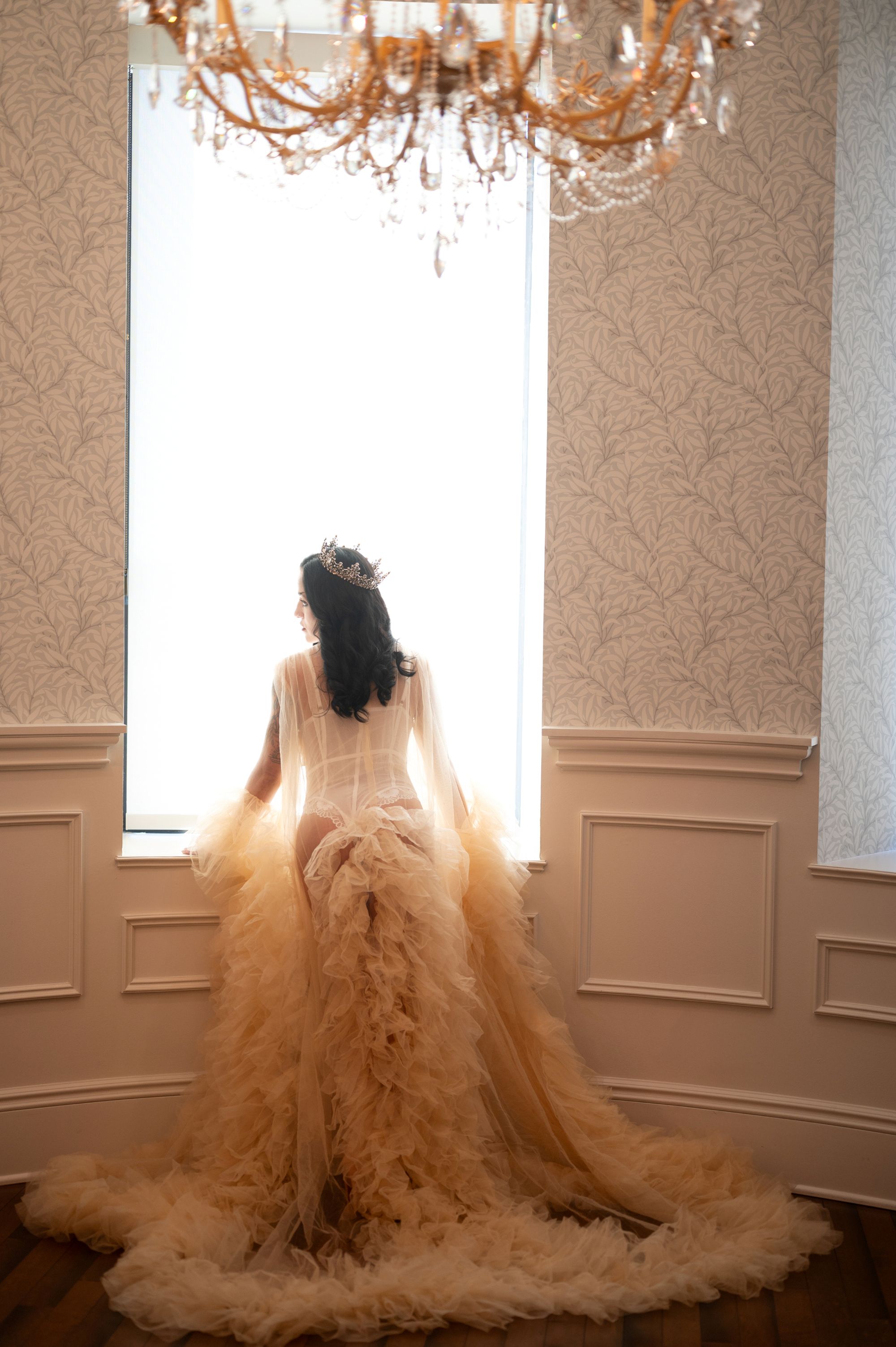 Regal editorial portrait of a woman in a flowing golden tulle gown and jeweled crown gazing out of a tall window beneath a chandelier.