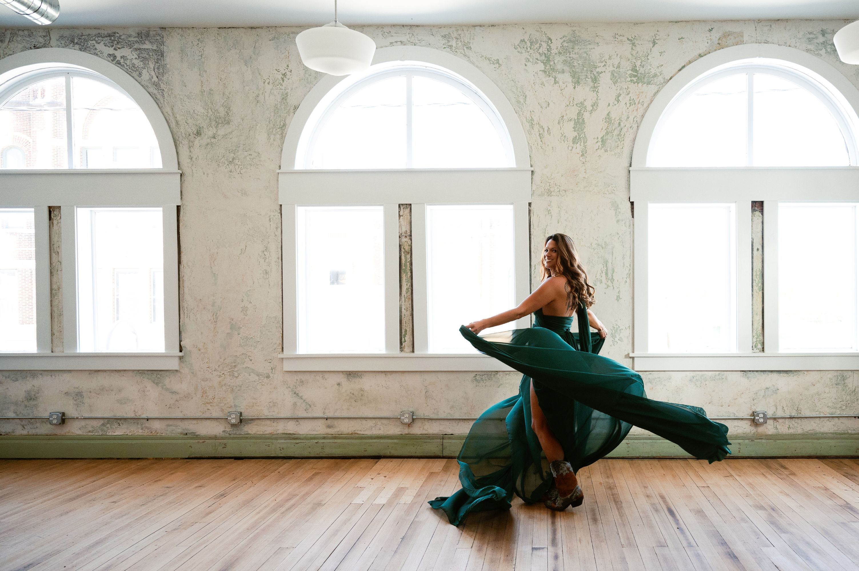 Woman in a flowing green gown twirls in a bright room with large arched windows and wooden floors.