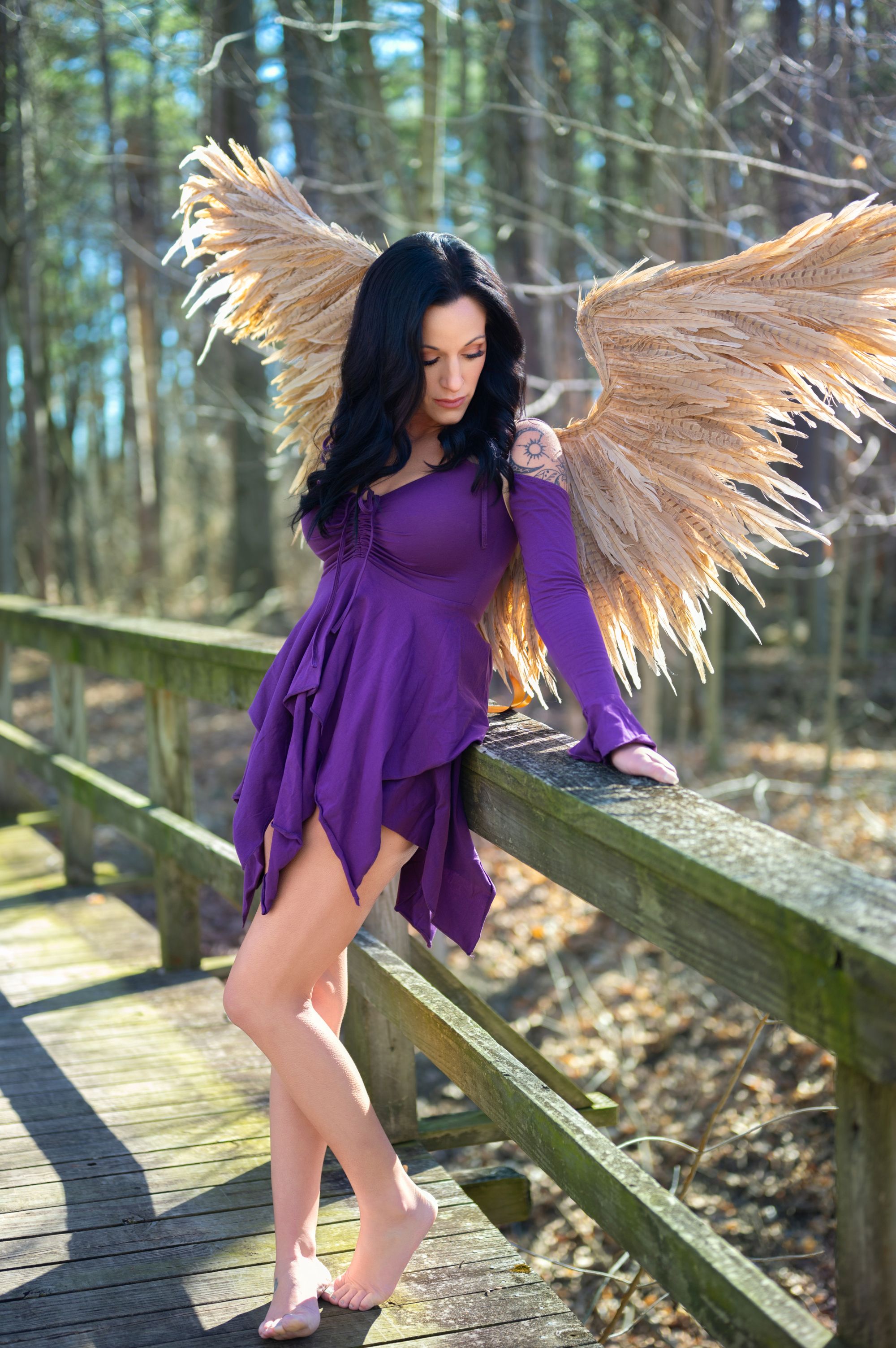 Fantasy-themed portrait of a woman with long black hair and feathered angel wings wearing a short purple dress on a wooden bridge in the forest.