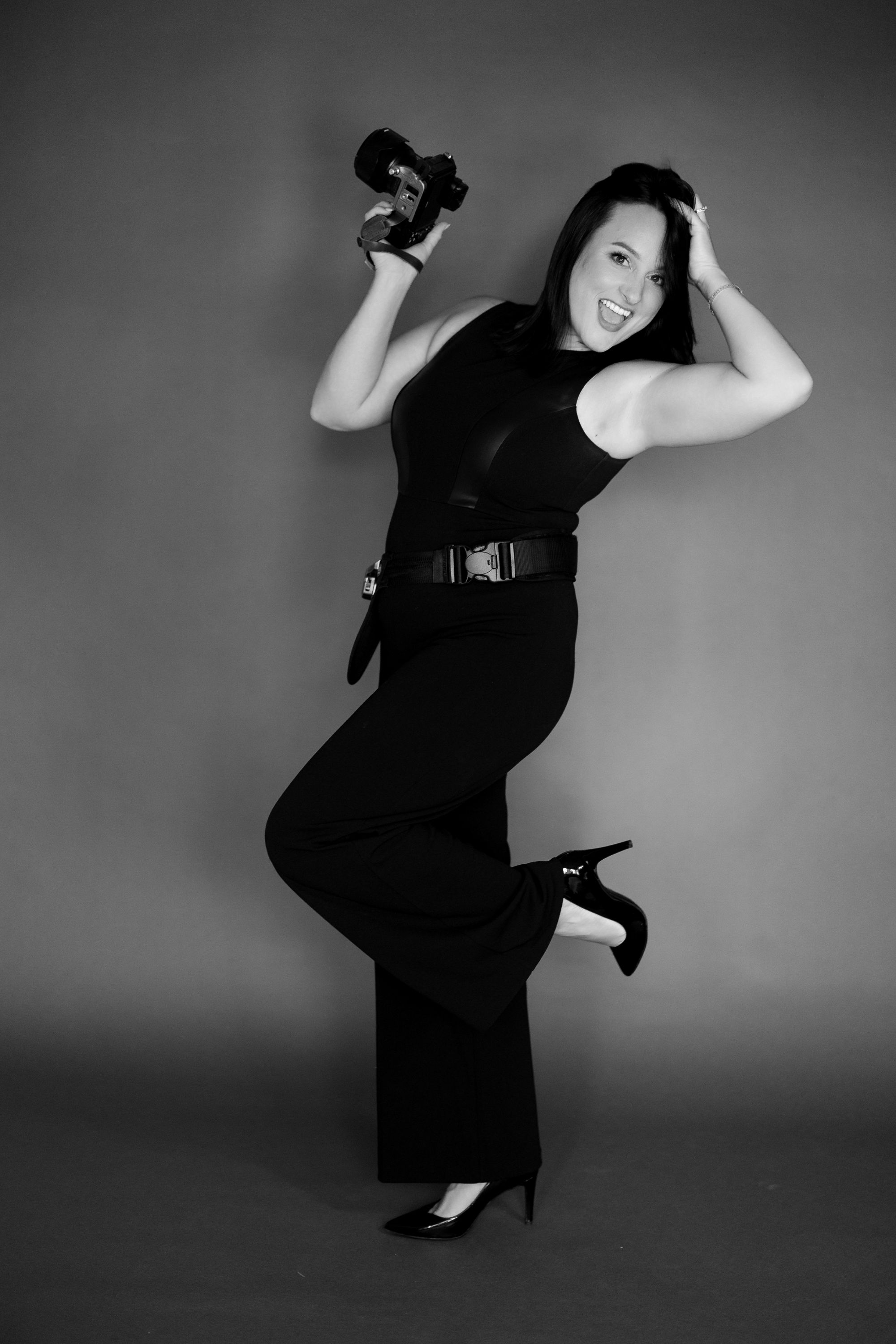 Playful portrait of a smiling woman in black outfit holding a camera in studio setting