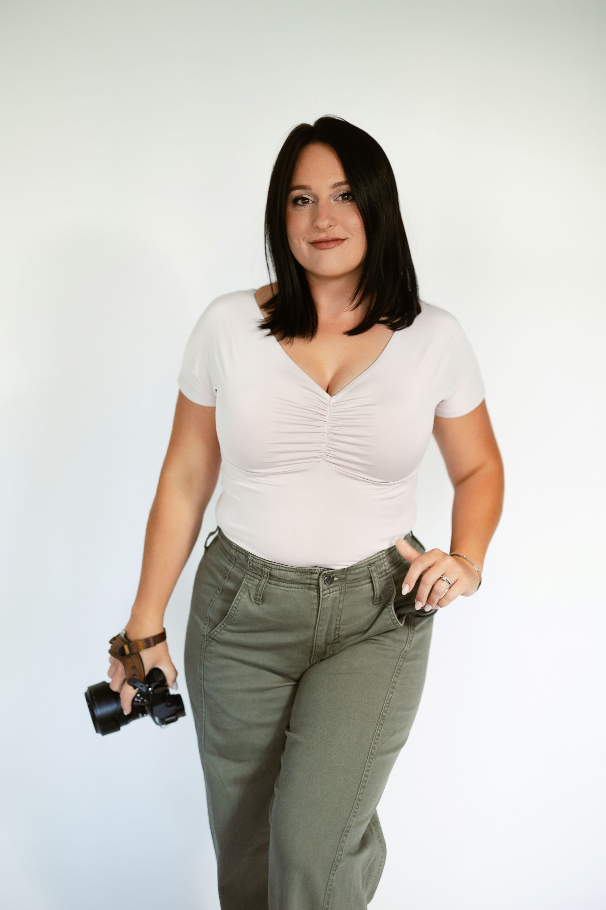 Confident female photographer in white top and green pants holding a camera against a white background