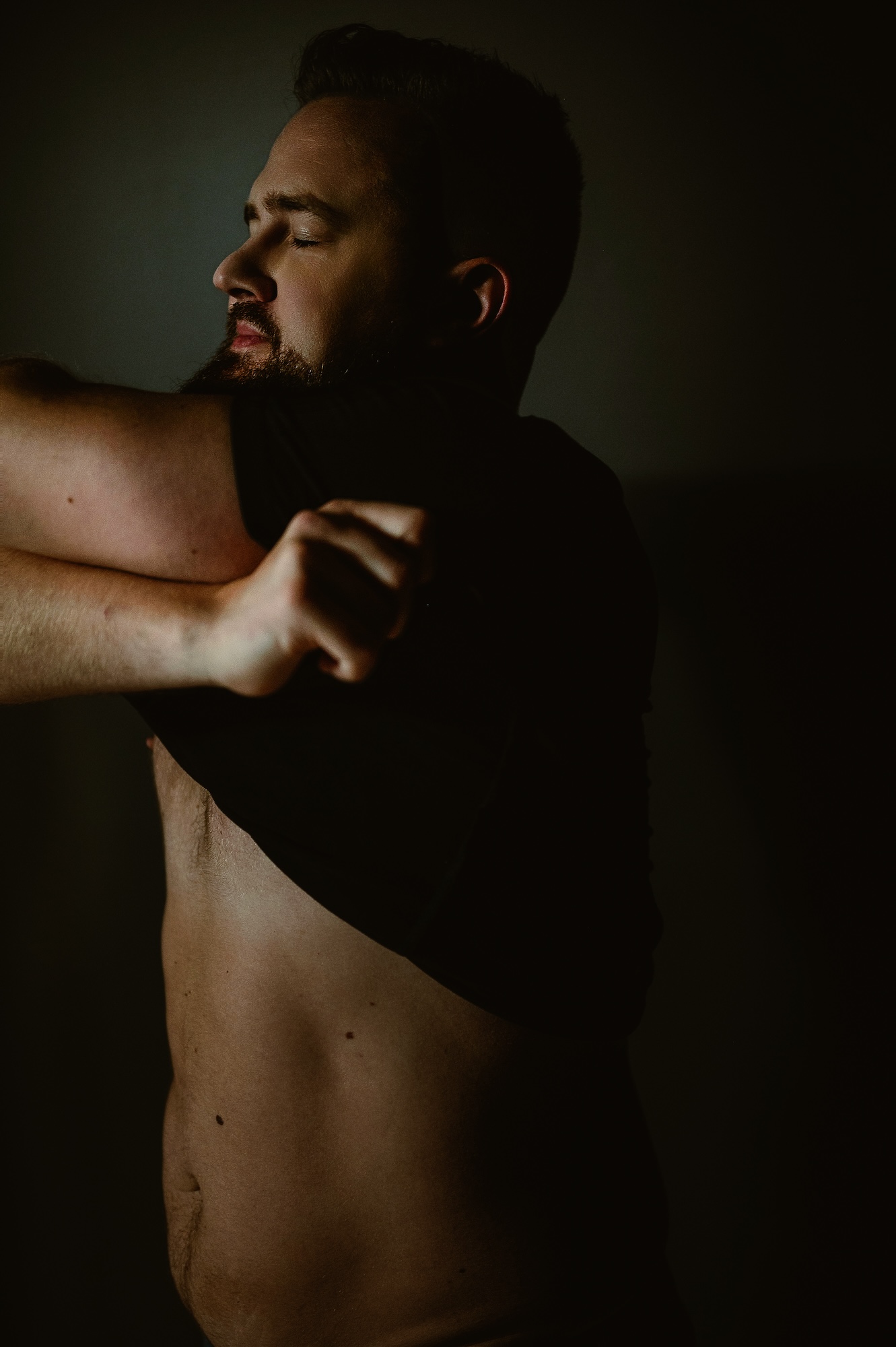 Artistic male boudoir photography of a man lifting his shirt with eyes closed, embracing self-confidence and vulnerability.