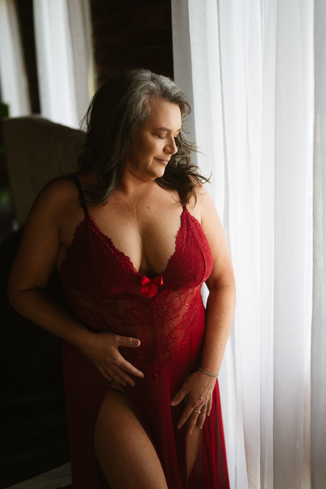 Empowering boudoir session of woman in red lace gown standing gracefully by window