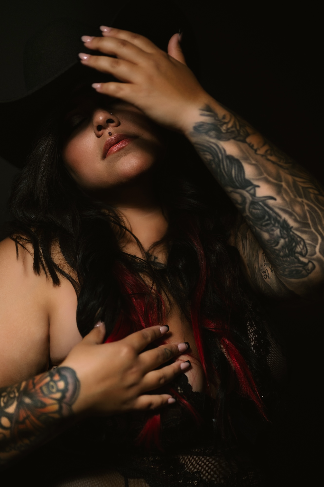 Moody boudoir photo of tattooed woman in black lingerie and cowboy hat with dramatic lighting