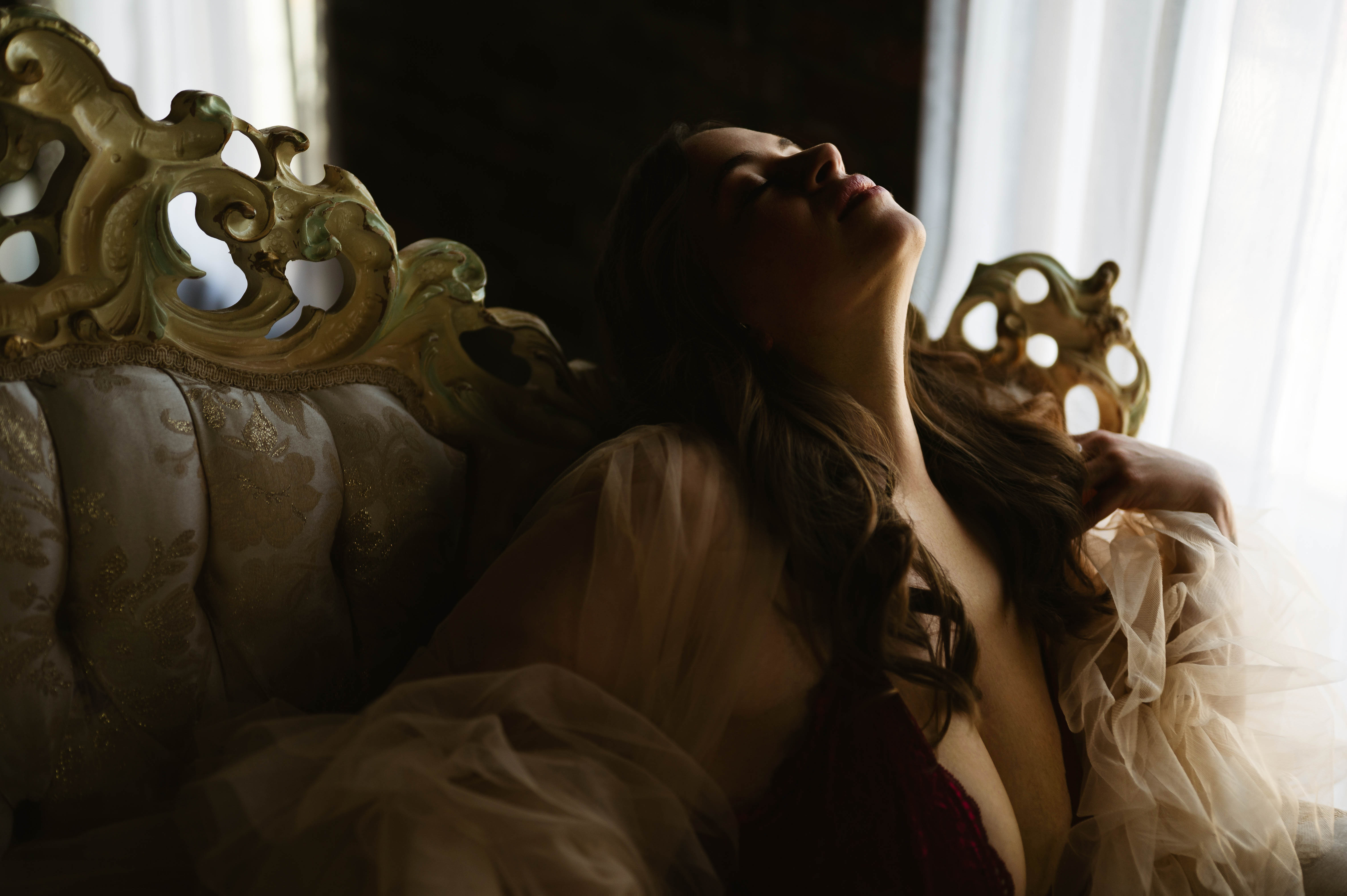 Woman in a sheer robe leans back on an ornate vintage sofa, illuminated by light streaming through a nearby window.