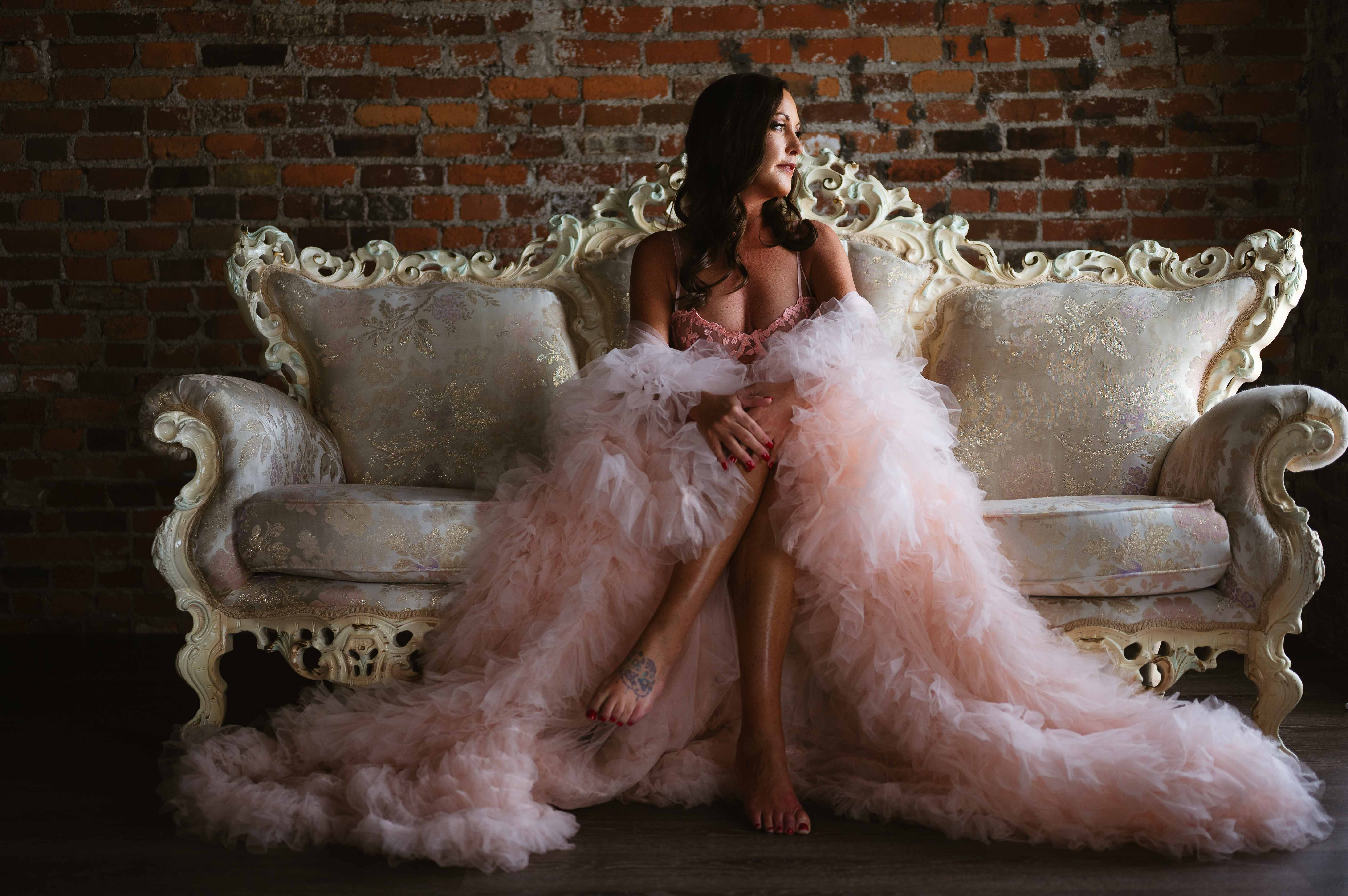 Woman draped in a flowing pink tulle robe sits on an ornate sofa against a brick wall, looking off to the side.