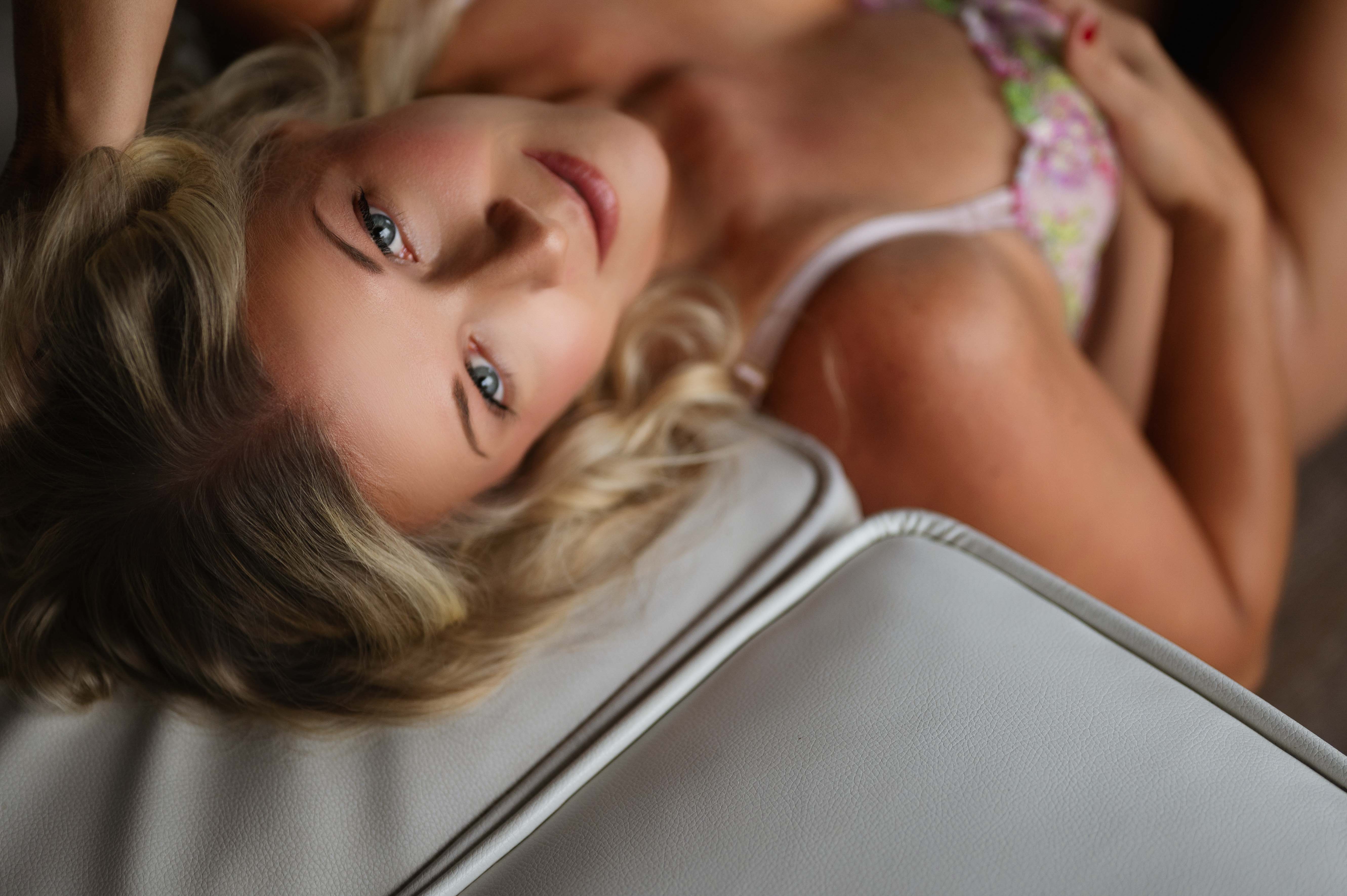 Woman in floral lingerie smiles gently while sitting on a white bed surrounded by soft light and curtains.