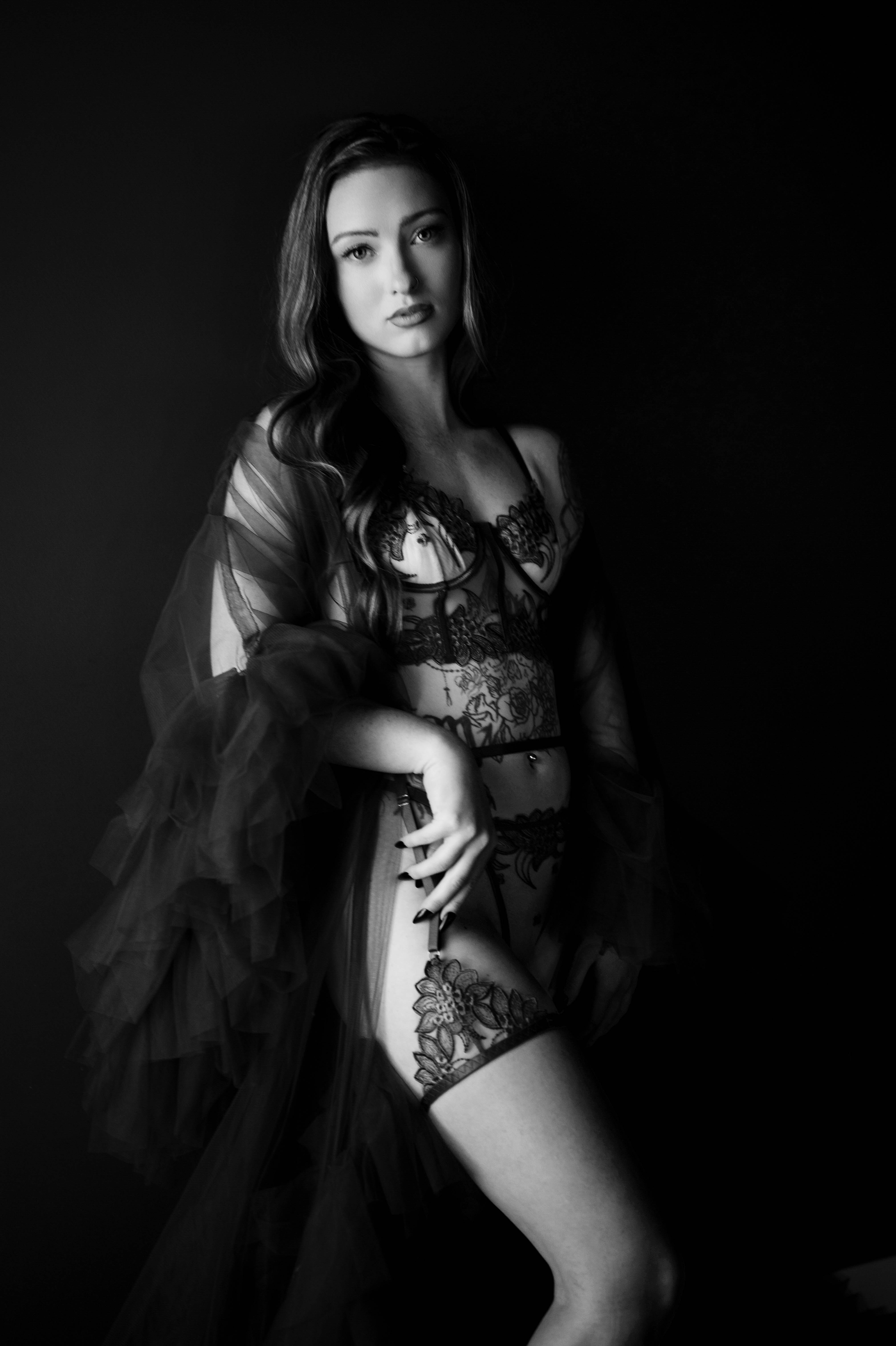 Woman with long wavy hair poses against a dark background in black lace lingerie and a sheer tulle robe, captured in black and white.