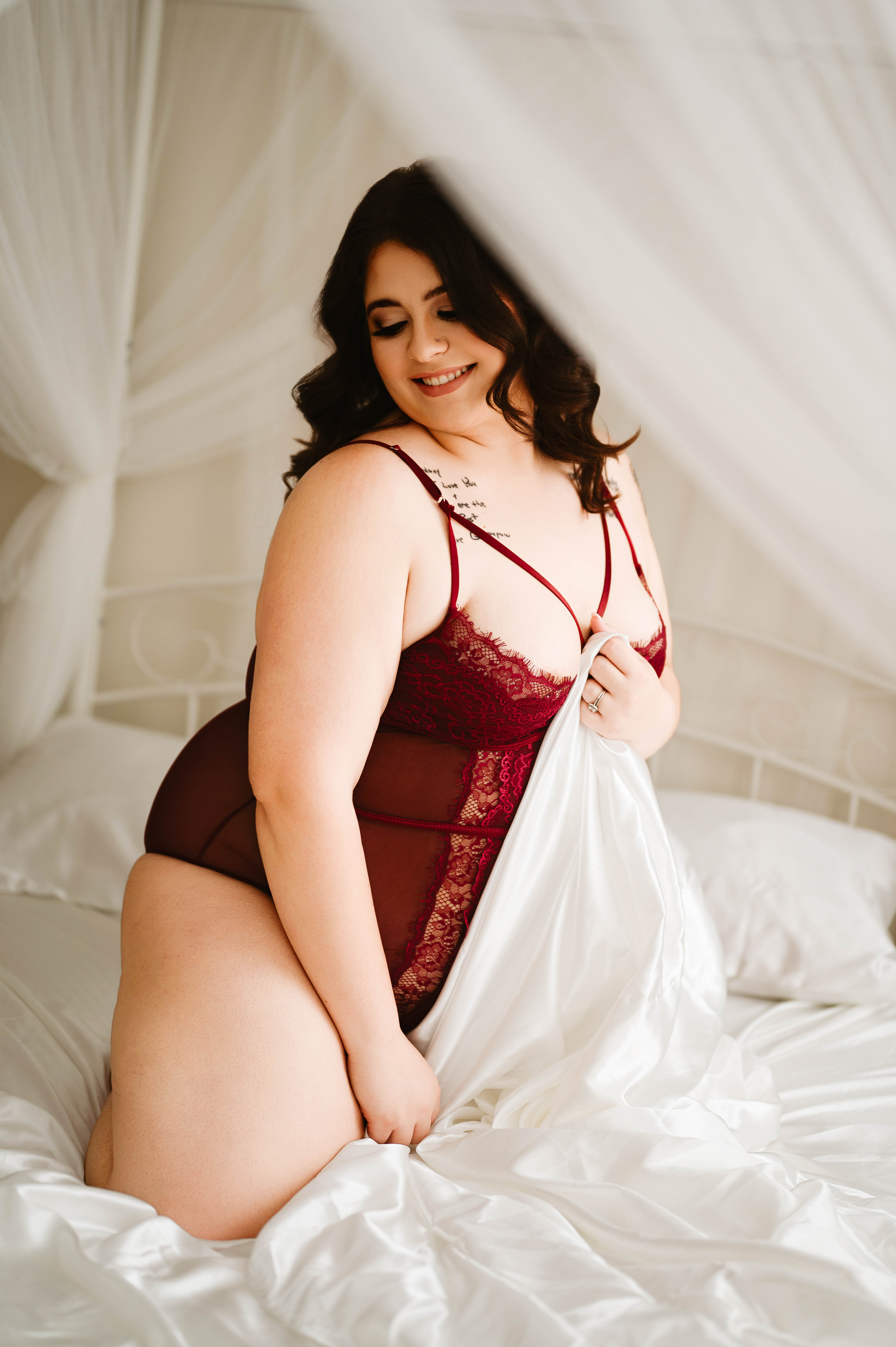 Woman with dark wavy hair kneels on a bed with white sheets, wearing burgundy lace lingerie and smiling softly.