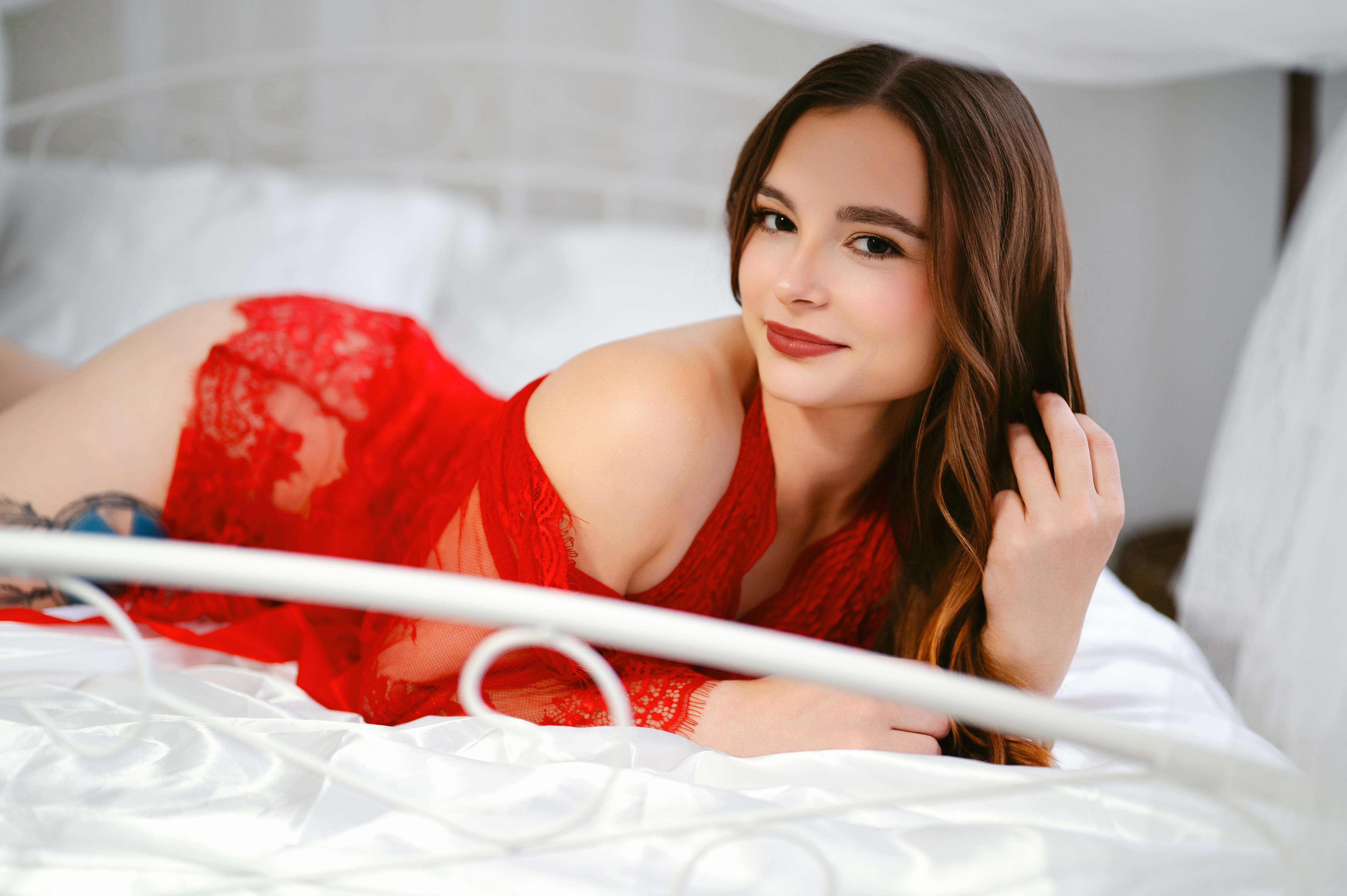 Woman in red lace lingerie lies on a bed, smiling at the camera in soft white light.