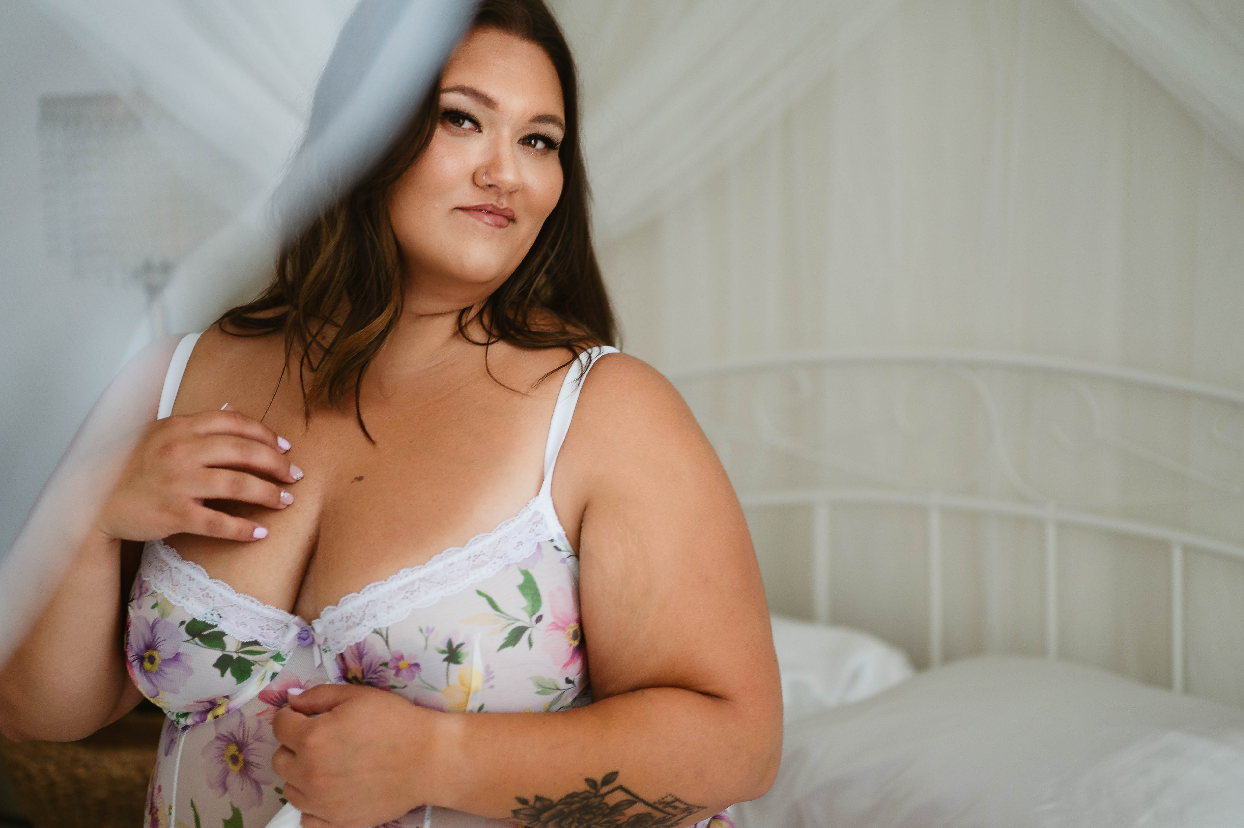Sultry boudoir photo of a tattooed woman in burgundy lingerie, posing playfully on a tufted white sofa.