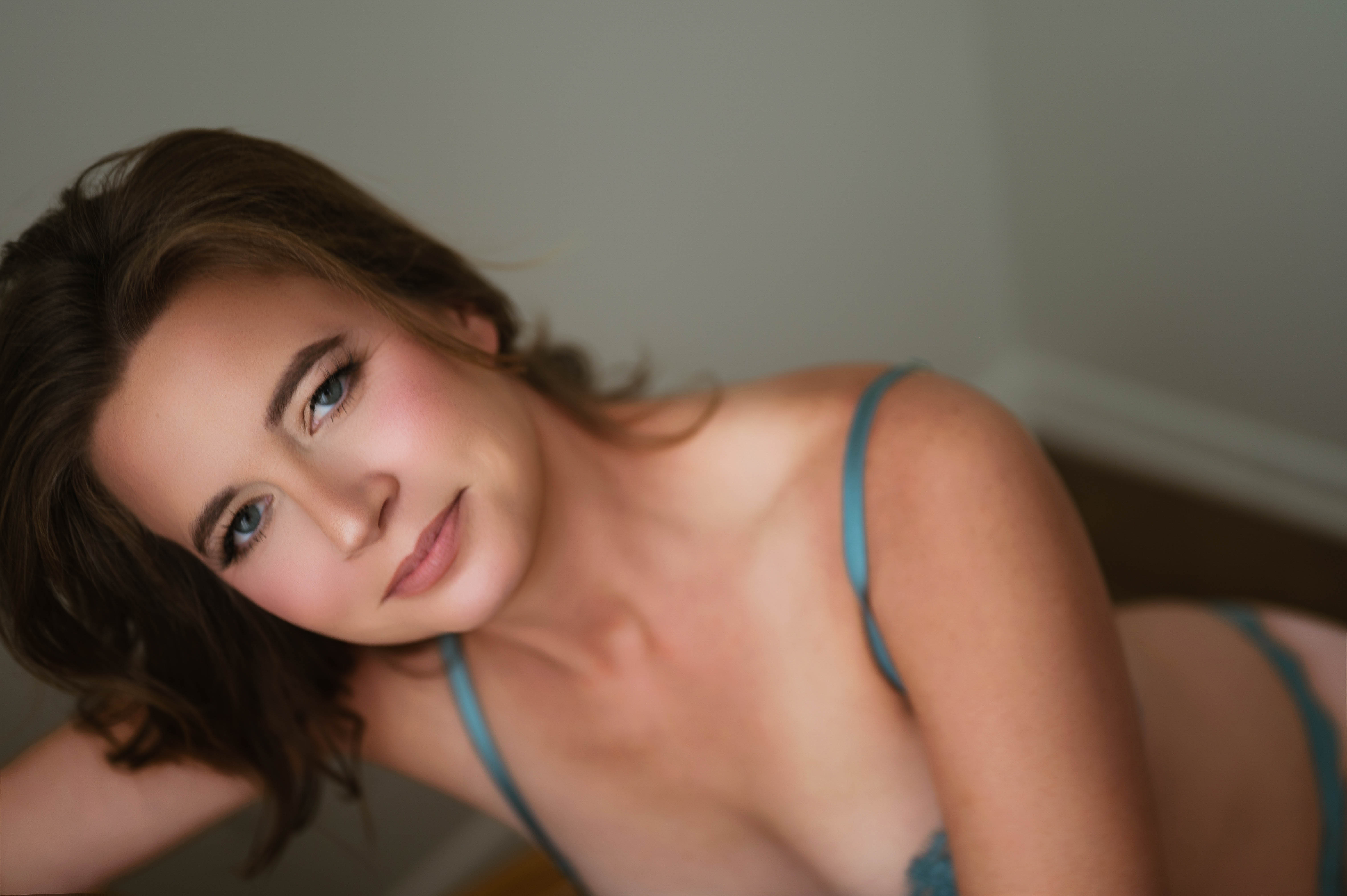 Woman in teal lingerie gazes at the camera, reclining softly against a neutral background.