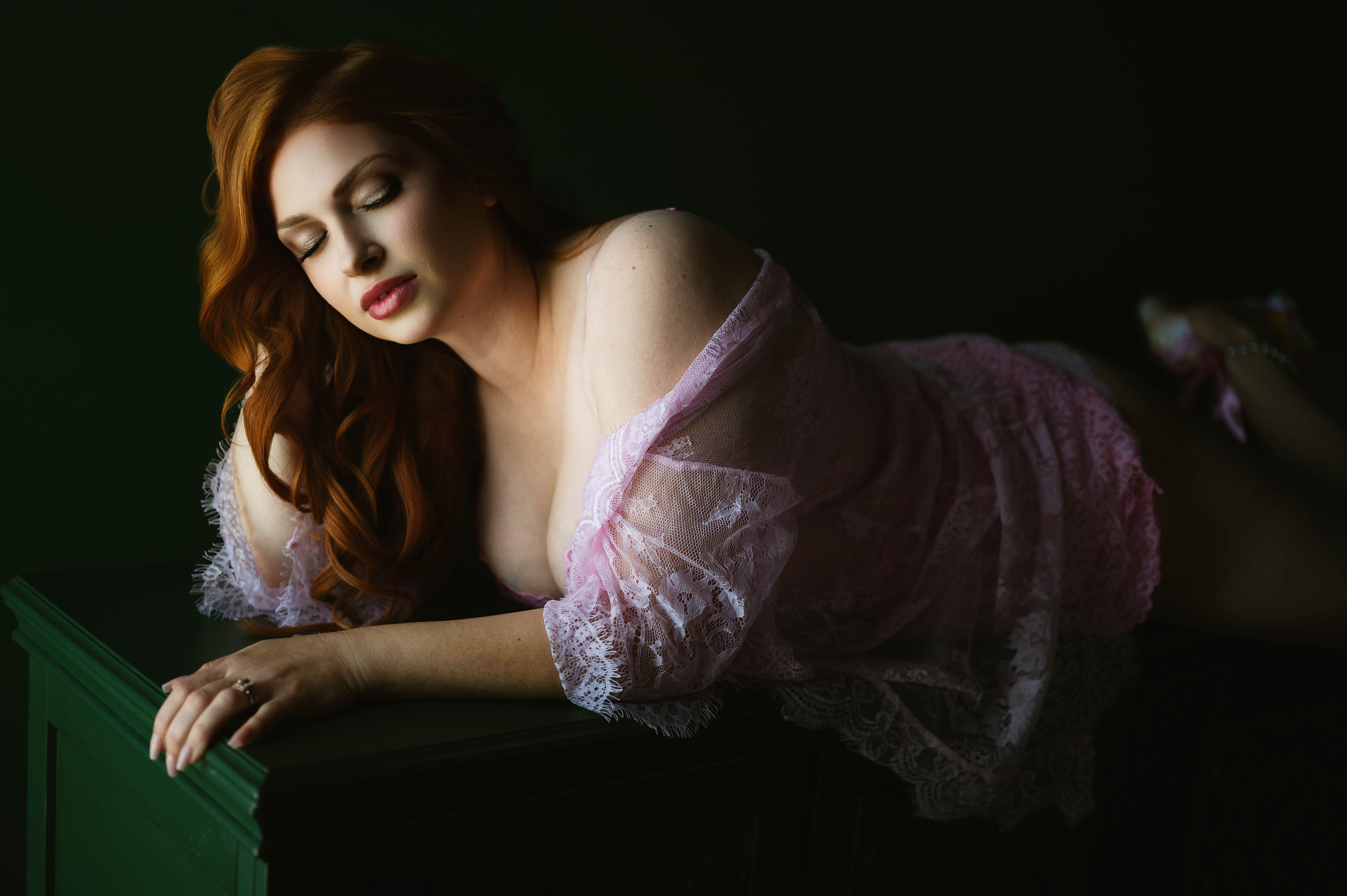 Woman with long wavy hair reclines on a green surface wearing a pink lace robe, softly lit in a relaxed pose.