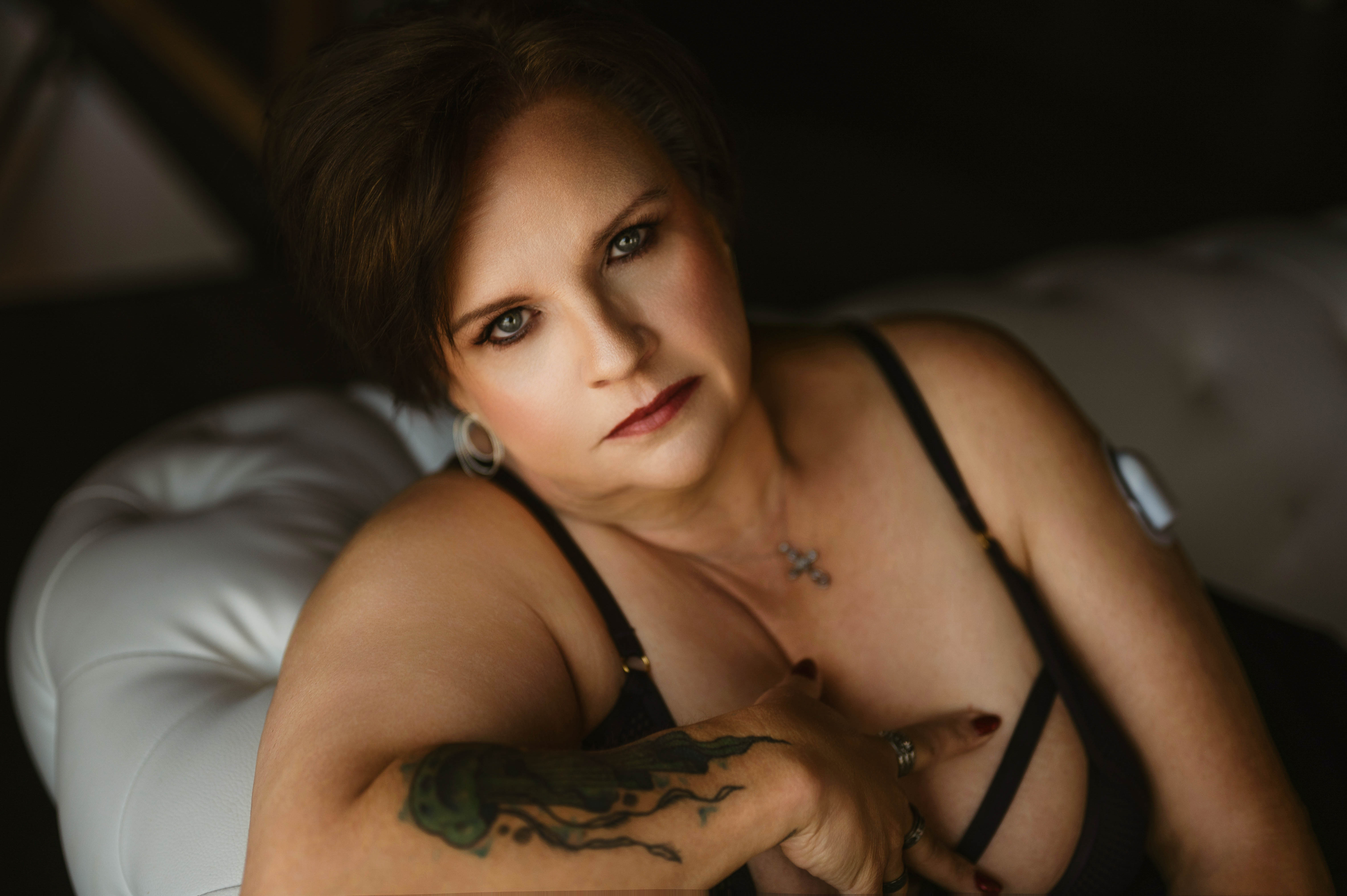 Woman with short hair and a tattoo on her arm gazes confidently at the camera while reclining on a white sofa in black lingerie.