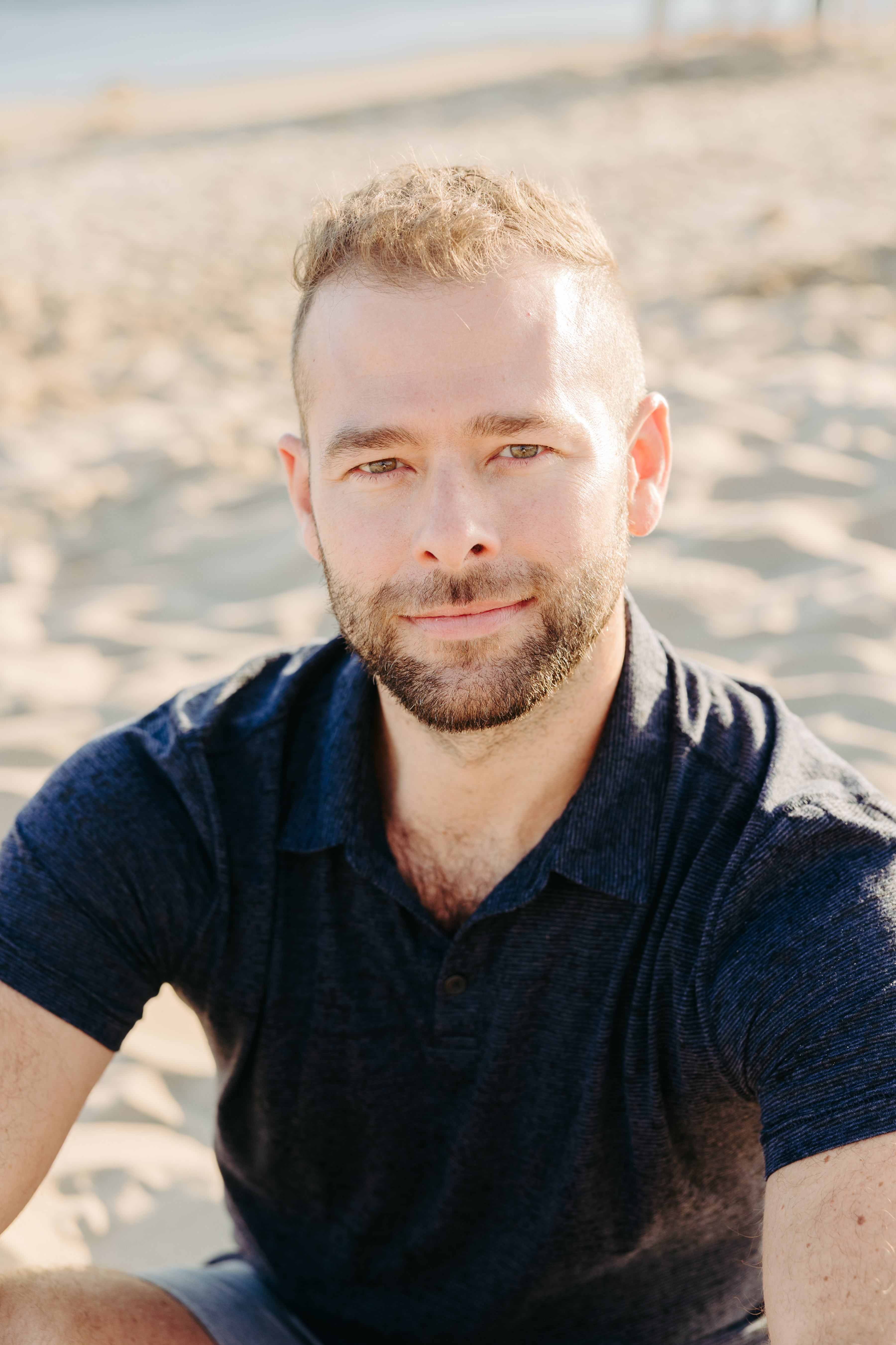 Jon Pinney seated on a sunlit beach, looking toward the camera with a gentle smile.
