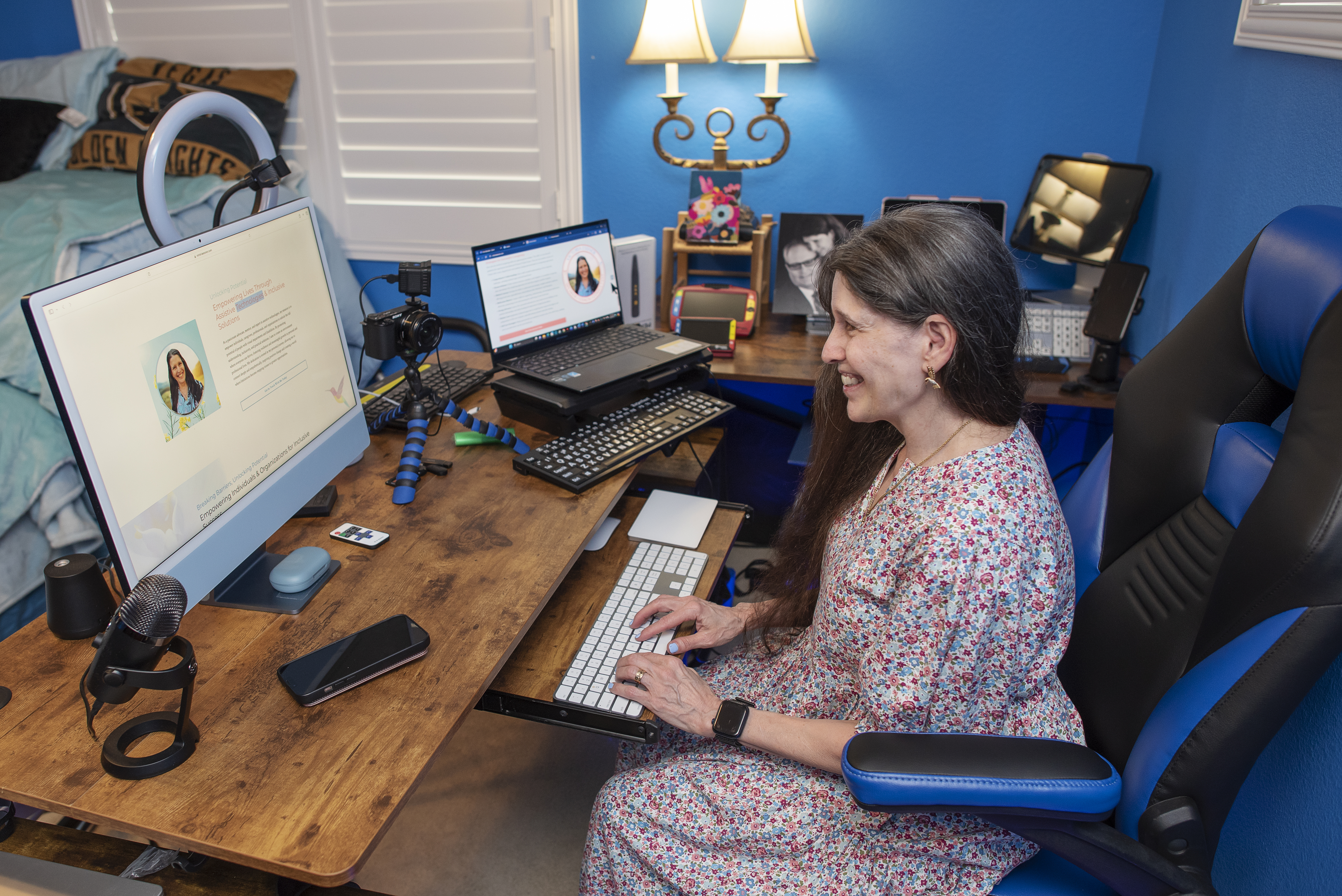 Victoria Essner smiles while working at her desk using multiple computers and assistive technology in her home office. The workspace includes a large keyboard, camera, microphone, and accessibility tools, reflecting her work as an Assistive Technology Coach and Job Accommodation Advisor. Victoria Essner smiles while working at her desk using multiple computers and assistive technology in her home office. The workspace includes a large keyboard, camera, microphone, and accessibility tools, reflecting her work as an Assistive Technology Coach and Job Accommodation Advisor.